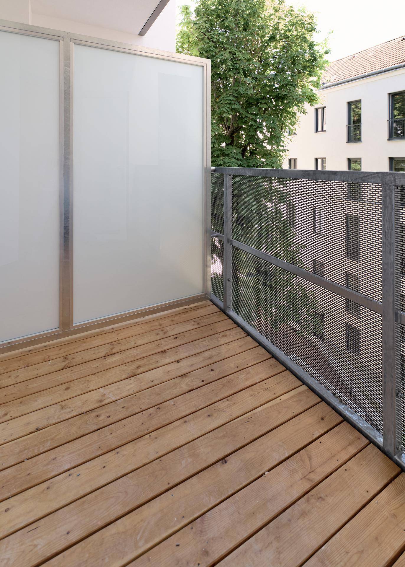 Balkon mit Holzdielenboden, Metallgeländer und Sichtschutz, umgeben von Bäumen.