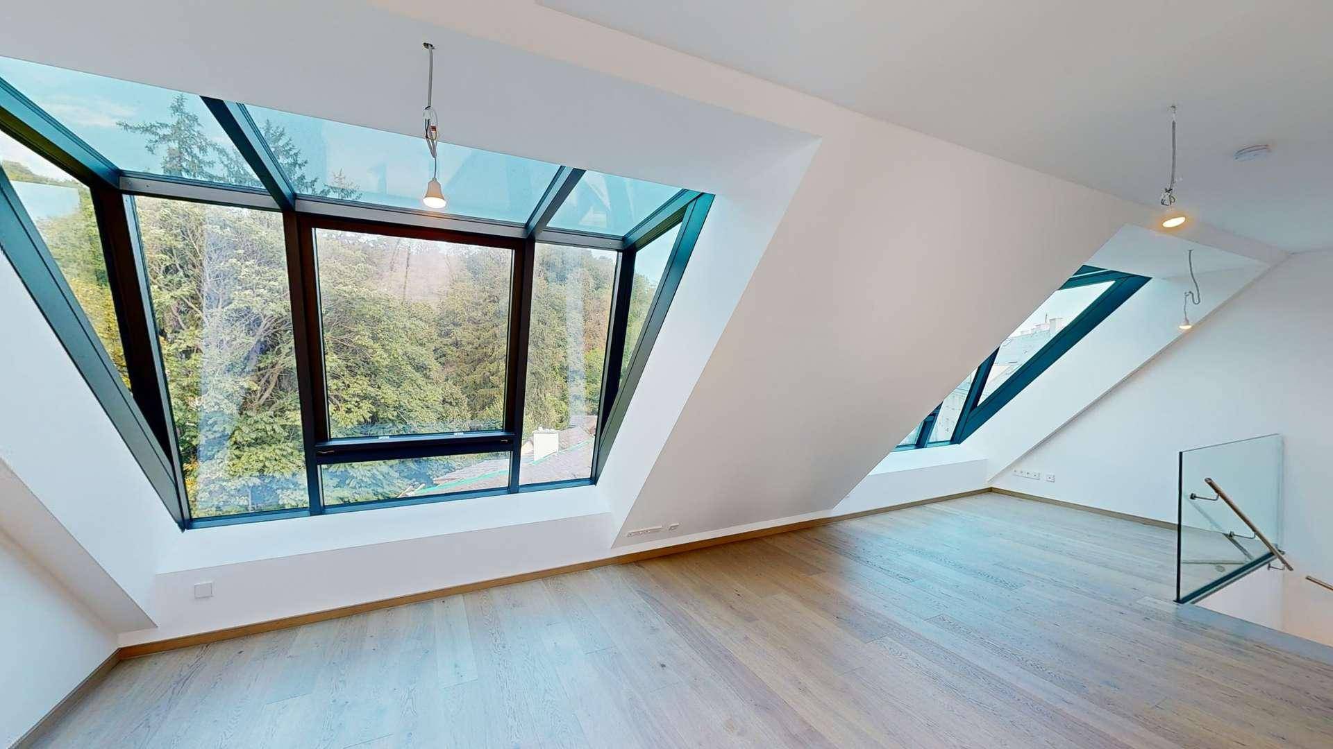 Lichtdurchfluteter Dachgeschossraum mit großen Fenstern, Holzfußboden und weitem Blick ins Grüne.
