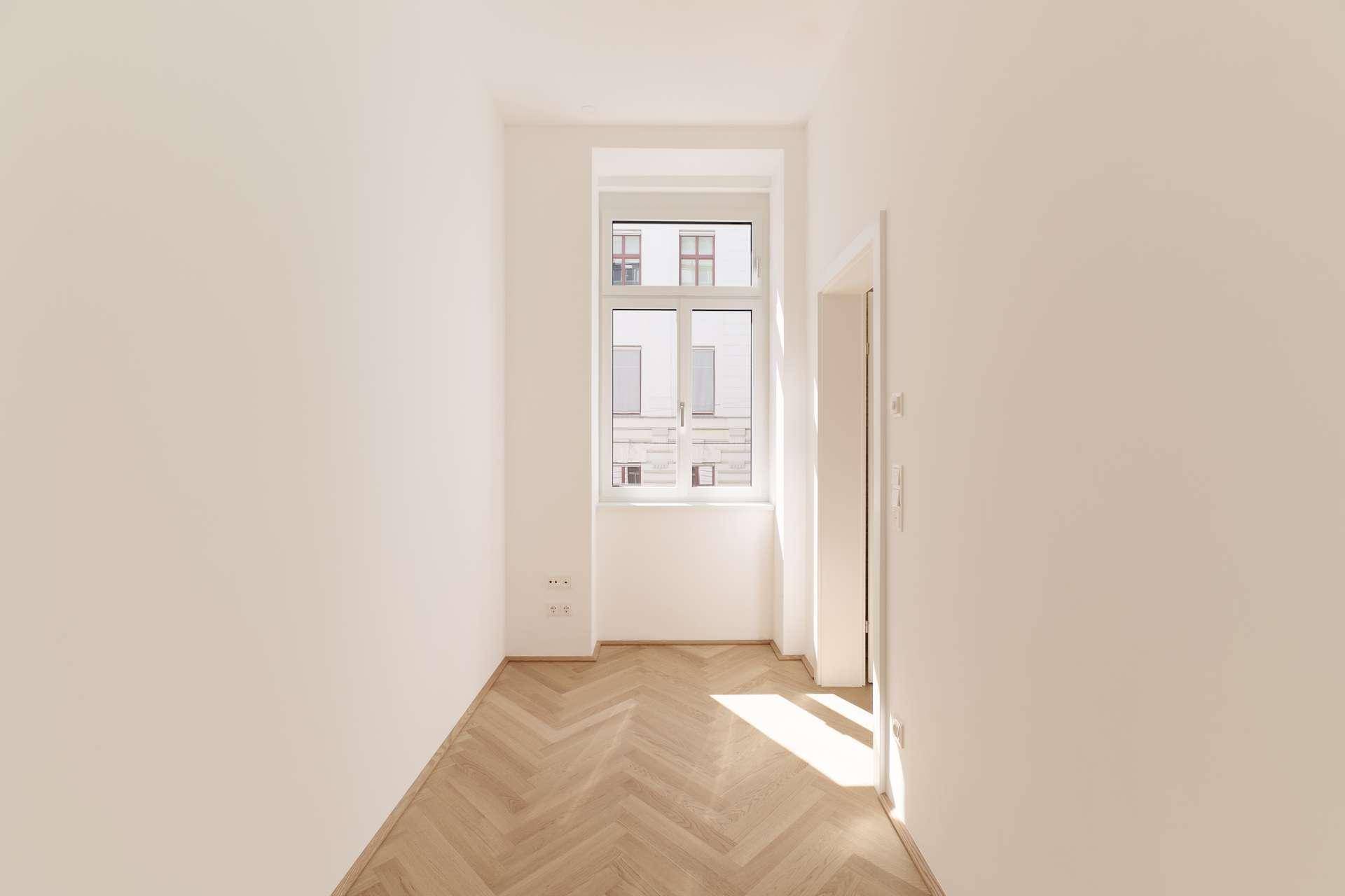 Helles Zimmer mit Fischgrätparkettboden und einem großen Fenster, das viel Tageslicht hereinlässt.