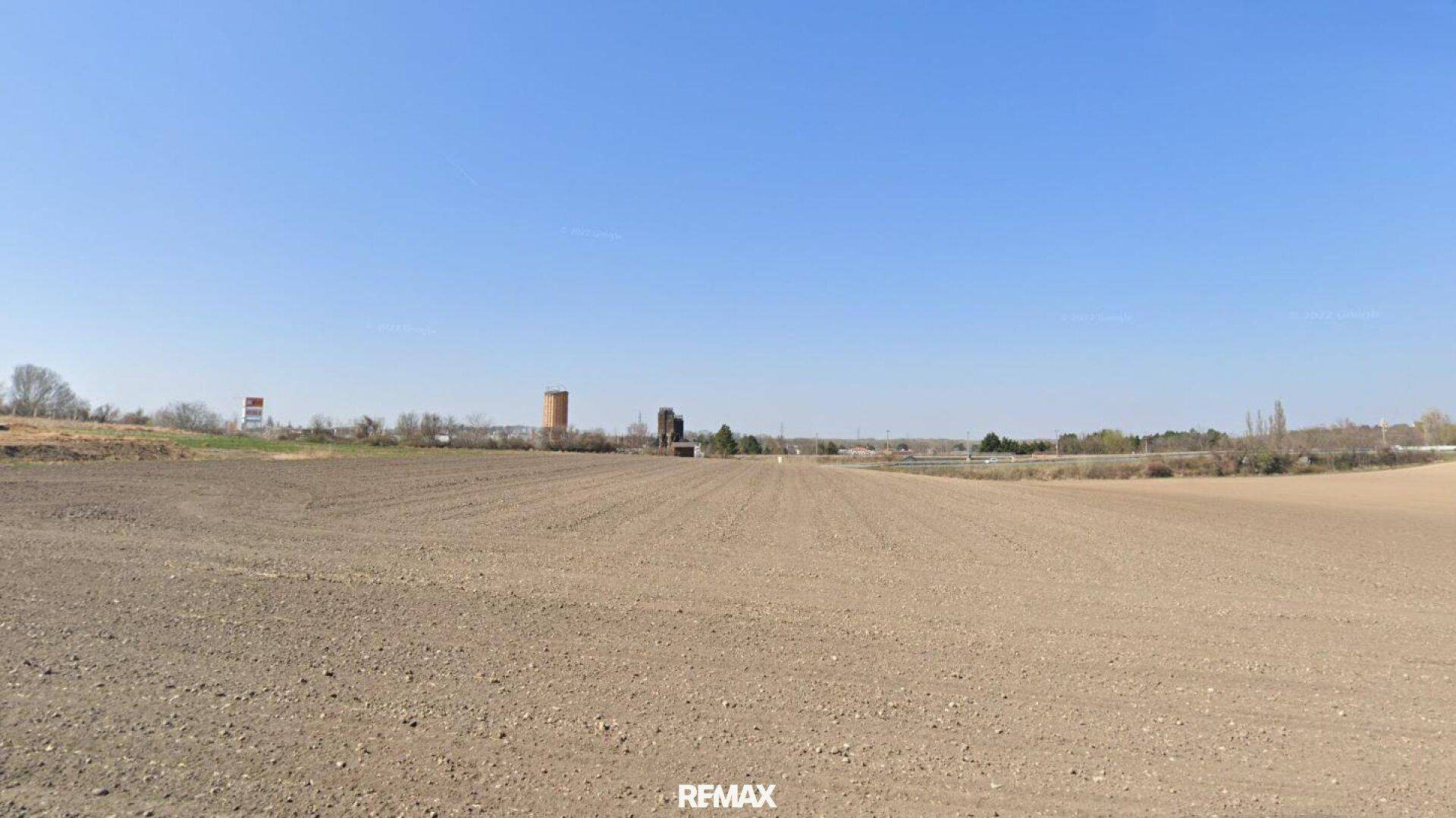 Weites, frisch gepflügtes Feld unter klarem blauem Himmel mit Blick auf entfernte Gebäude.