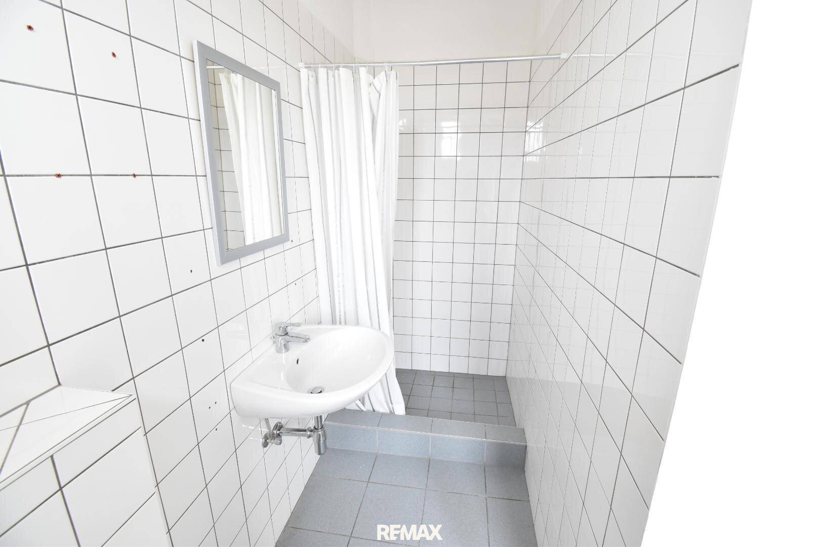 Einfaches Badezimmer mit weißen Fliesen, Waschbecken und Duschbereich mit Vorhang.