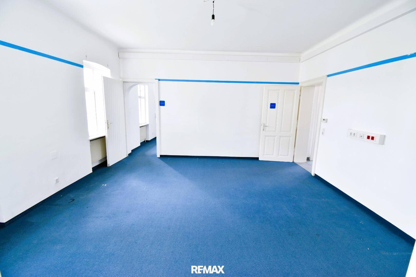 Heller, leerer Raum mit blauem Teppichboden und mehreren Türen zu angrenzenden Bereichen.