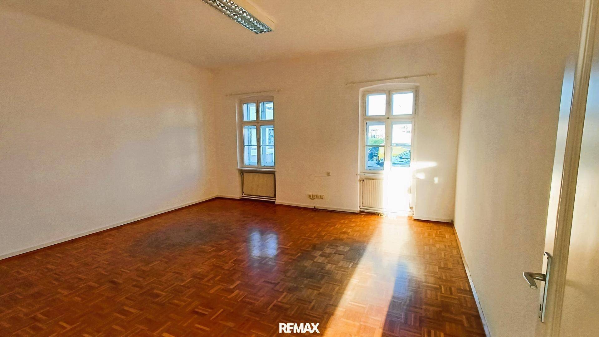 Heller Raum mit Holzparkettboden, zwei großen Fenstern und viel Sonneneinstrahlung.