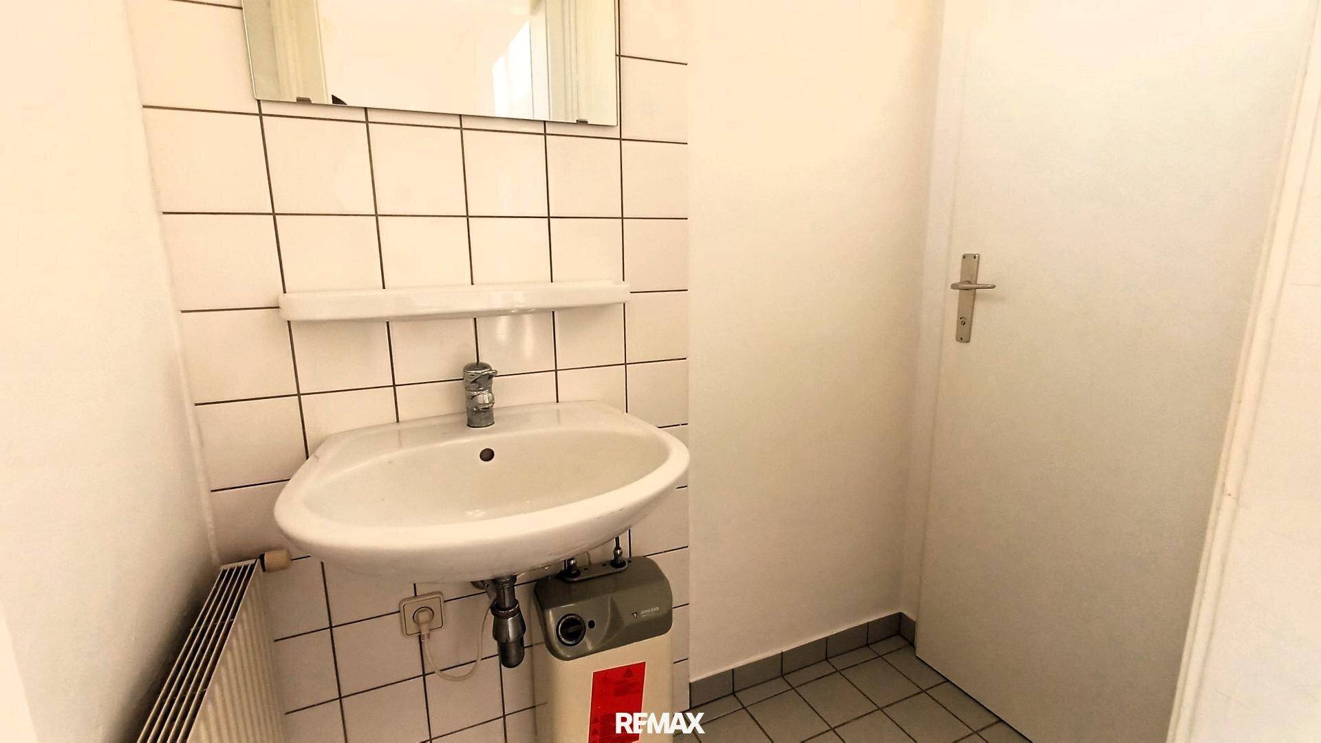 Badezimmer mit weißen Fliesen, Waschbecken, Heizkörper und einem kleinen Warmwasserbereiter.