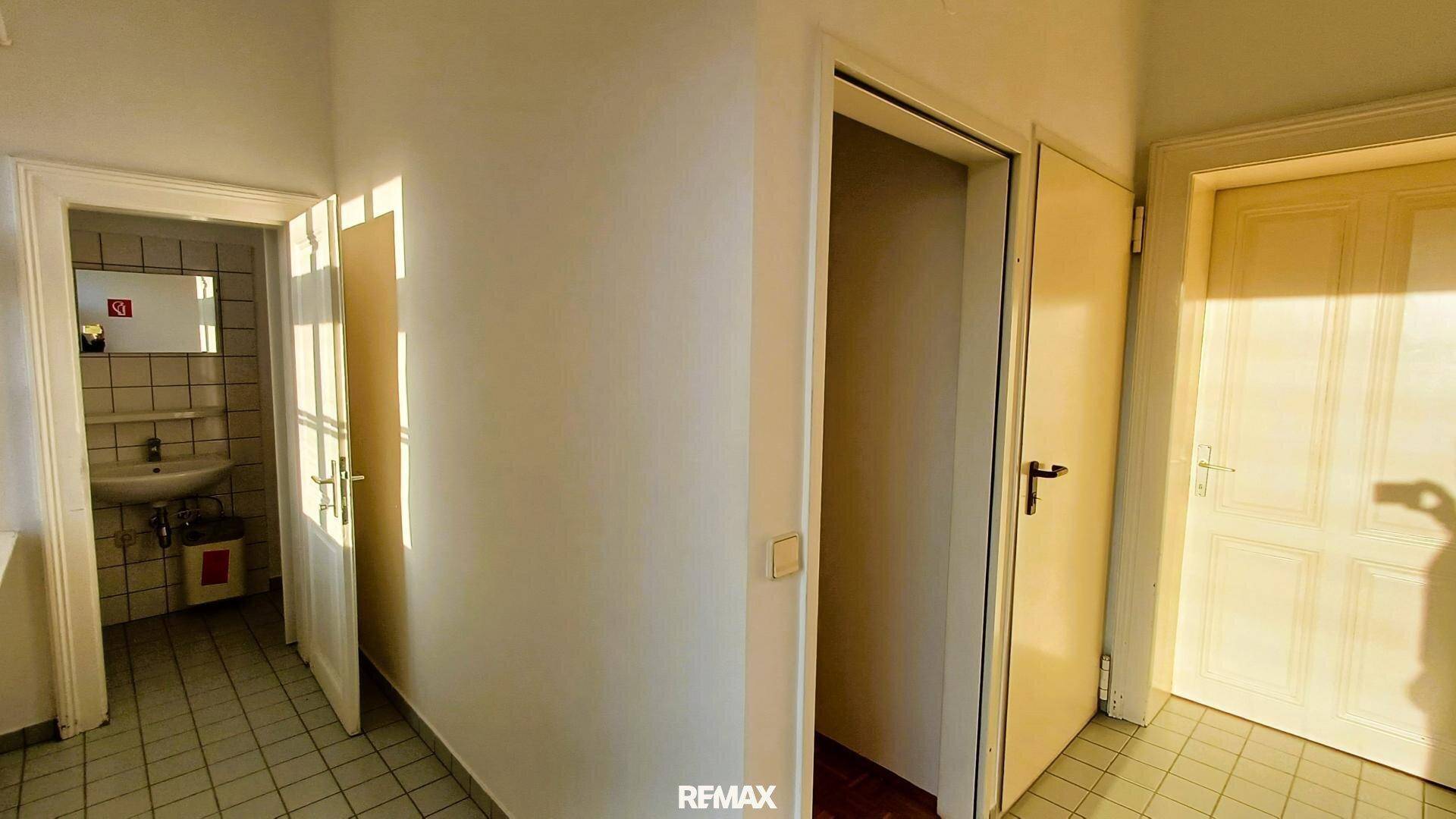Heller Flur mit Fliesenboden und Blick in ein Badezimmer mit Waschbecken und Spiegel.