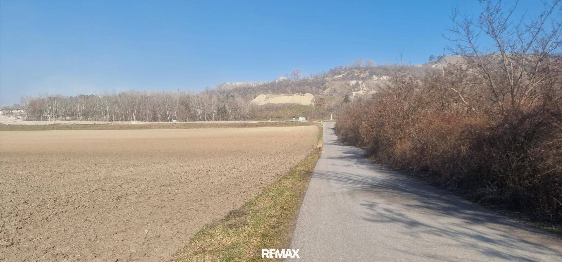 Ländliche Umgebung mit einem Feldweg, trockenen Feldern und bewaldeten Hügeln im Hintergrund unter blauem Himmel.