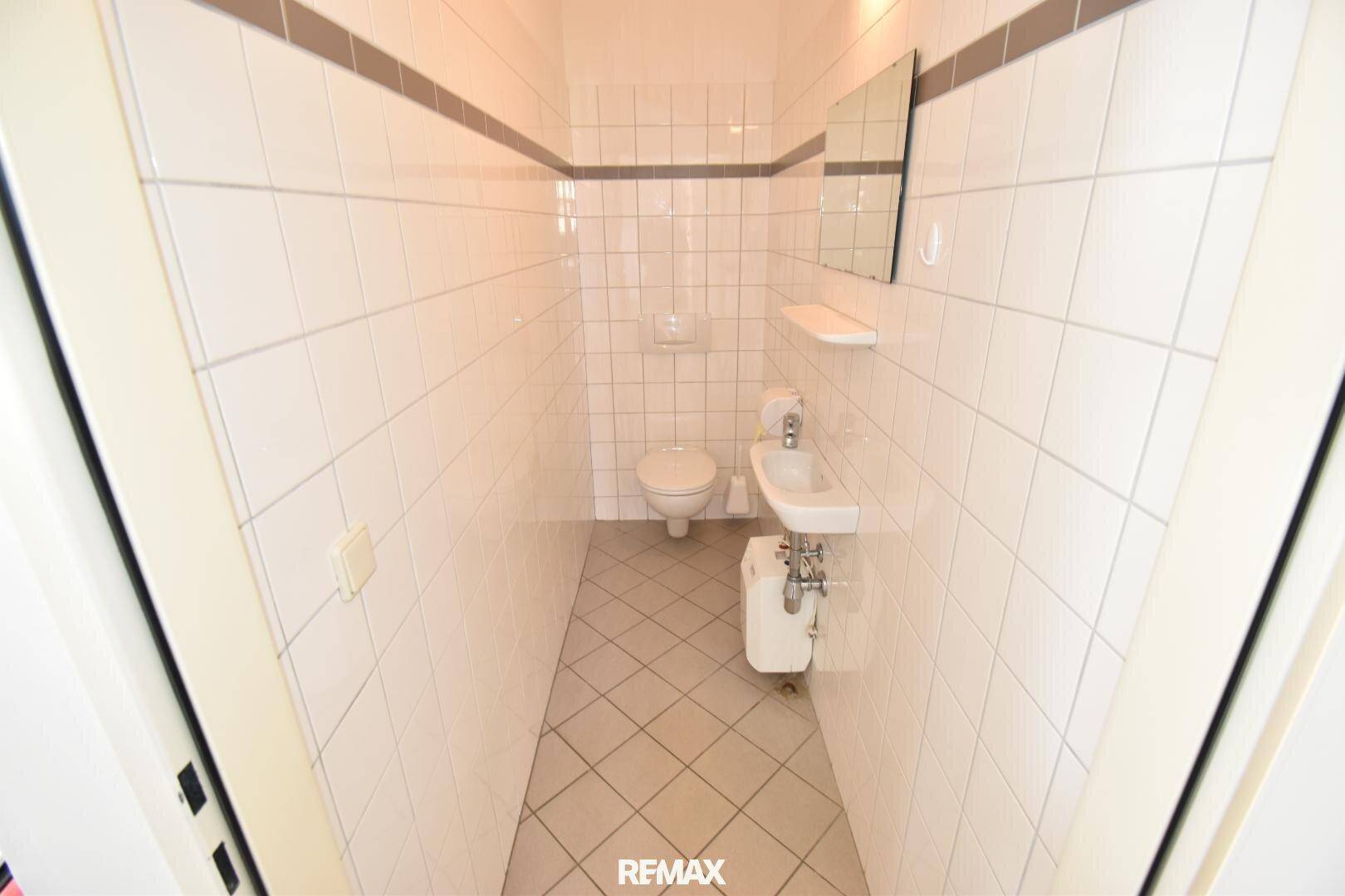 Schmale Toilette mit weißen Fliesen an Wänden und Boden, ausgestattet mit WC und kleinem Waschbecken.