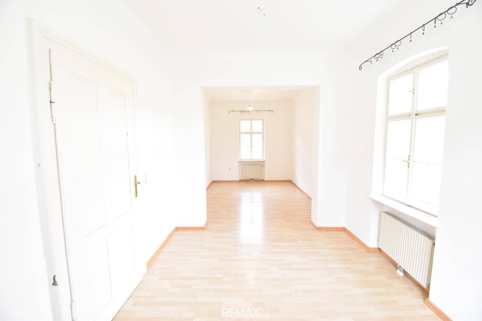 Leerer Raum mit hellem Laminatboden, weißen Wänden, einem Fenster und einer weißen Tür.