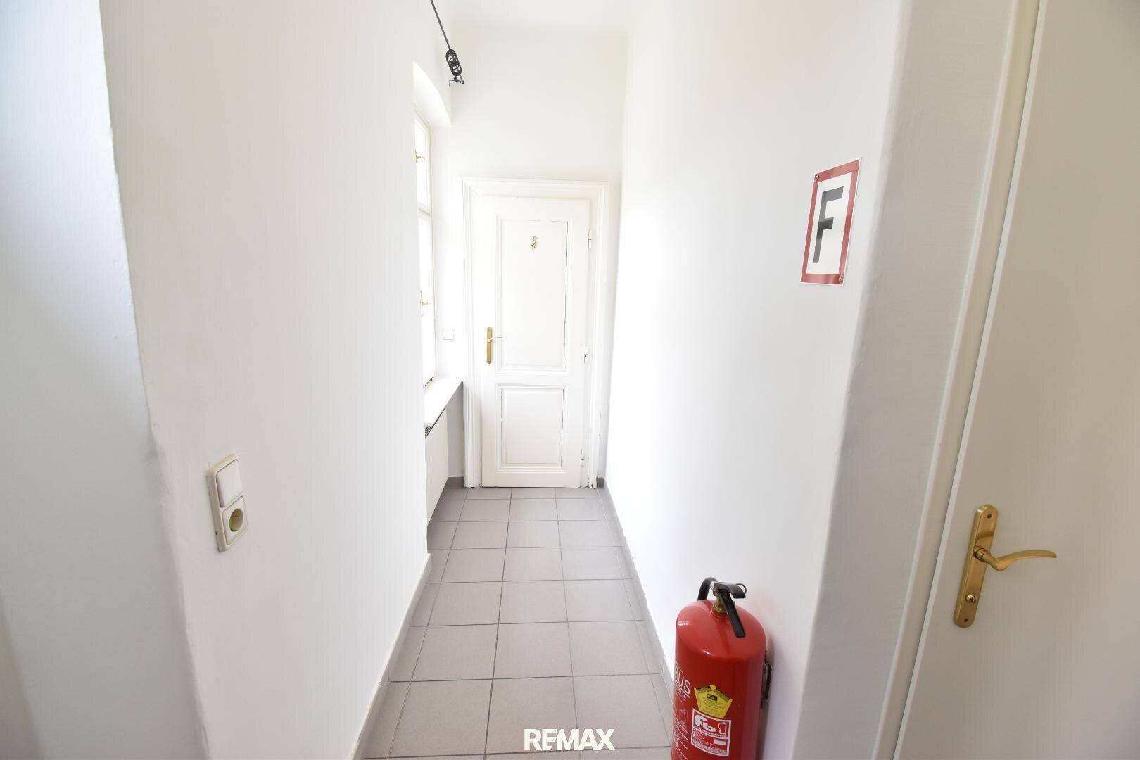 Flur mit grauem Fliesenboden, weißen Wänden, einem Fenster und einem Feuerlöscher.