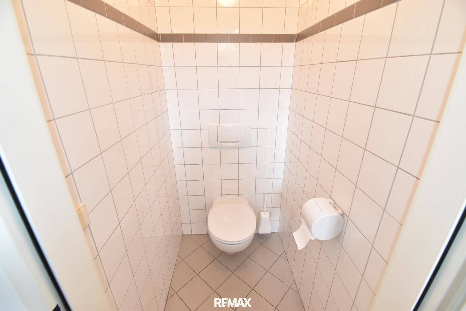Kleine Toilettenkabine mit weißen Fliesen an Wänden und Boden, ausgestattet mit einer Toilette.