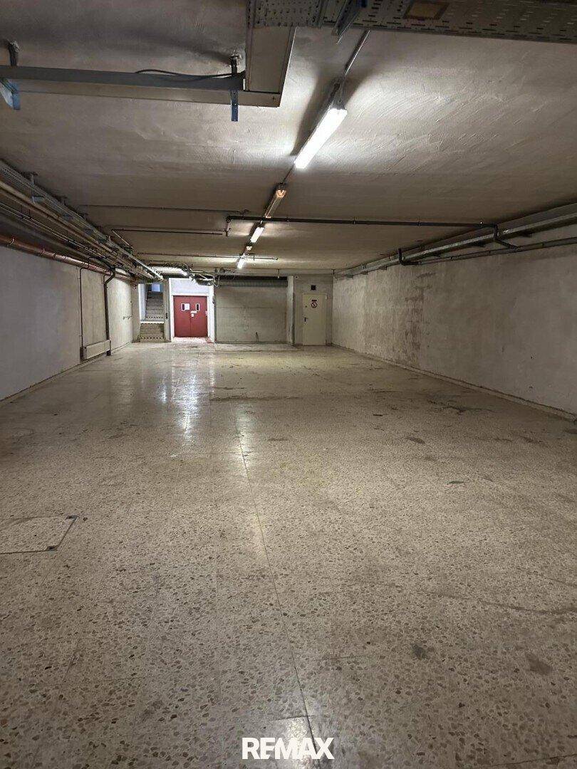 Geräumiger Kellerraum mit Betonboden, sichtbaren Leitungen an der Decke und einer roten Tür im Hintergrund.