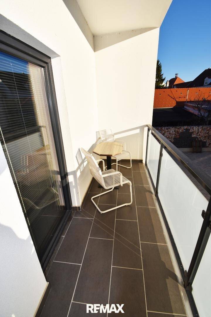Sonniger Balkon mit Fliesenboden, Glasgeländer und Sitzgelegenheiten.