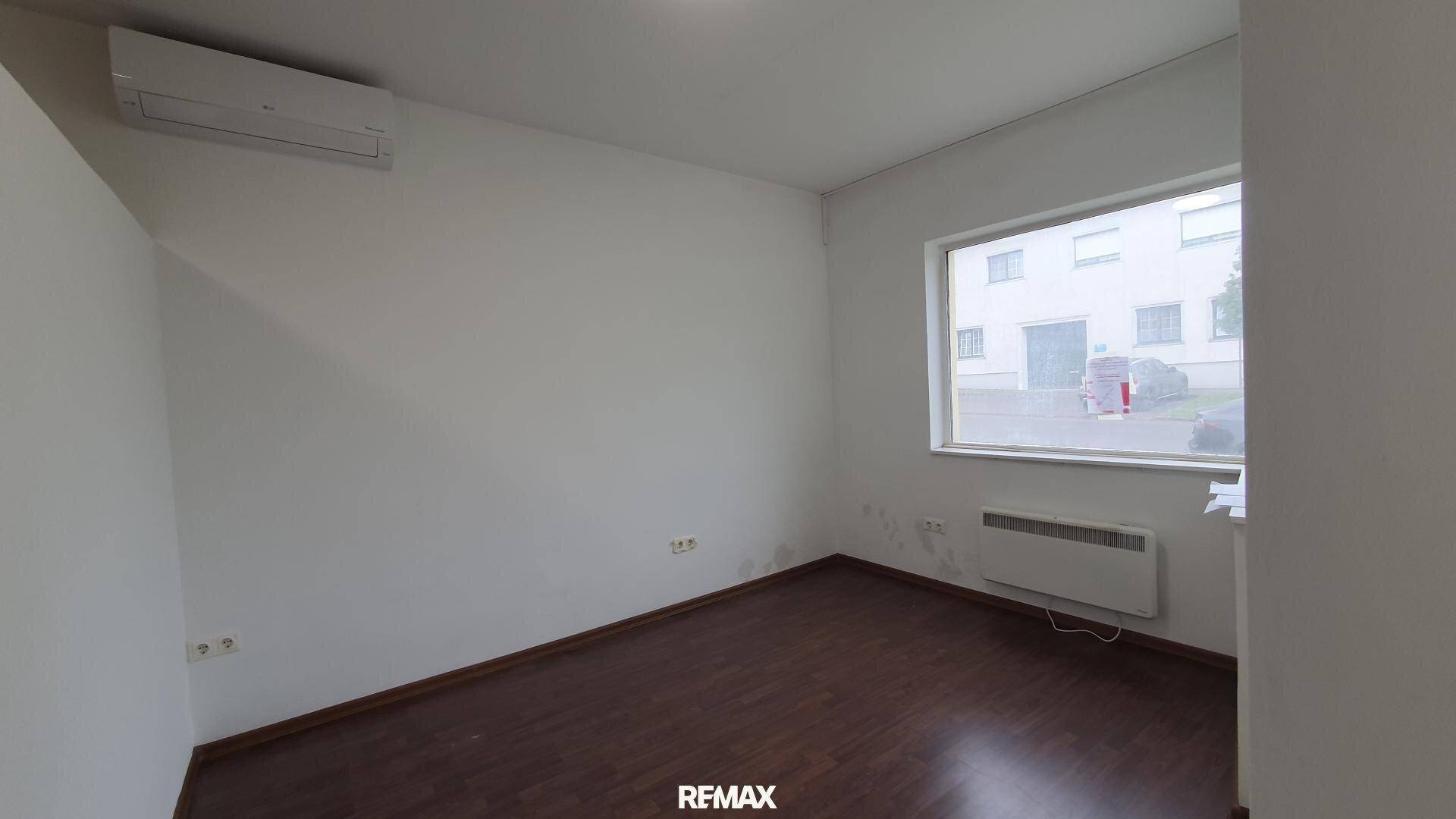 Heller Büroraum mit dunklem Laminatboden, weiß gestrichenen Wänden und einem großen Fenster.