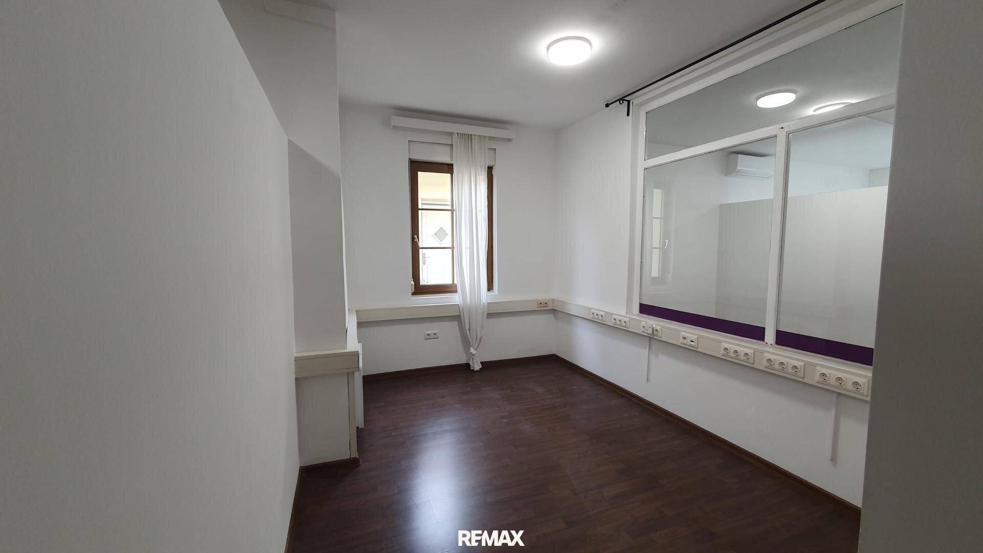Büroraum mit Laminatboden, einem Fenster und einer internen Glaswand, die Licht in den Nebenraum lässt.