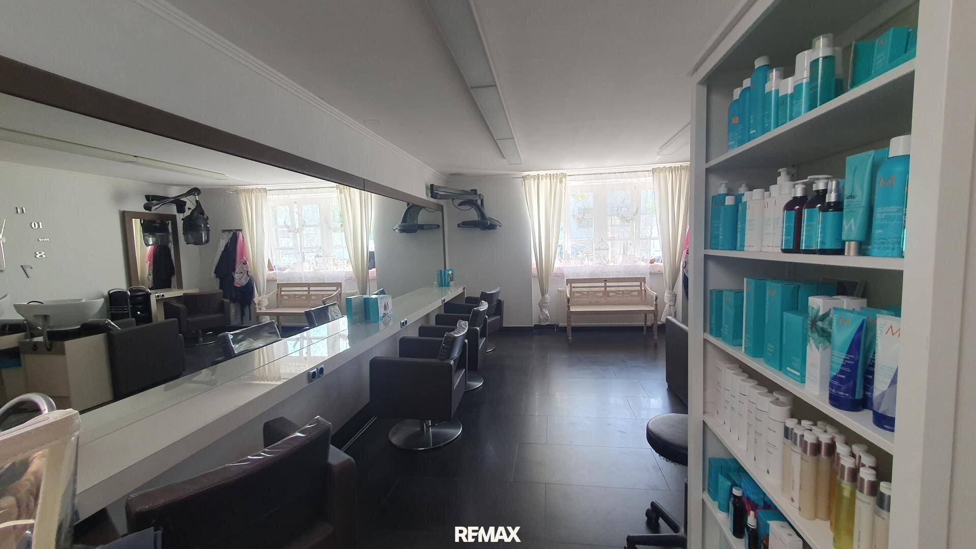 Langer Friseursalon mit vielen Styling-Stühlen, einem durchgehenden Spiegel und Fenstern mit Vorhängen.