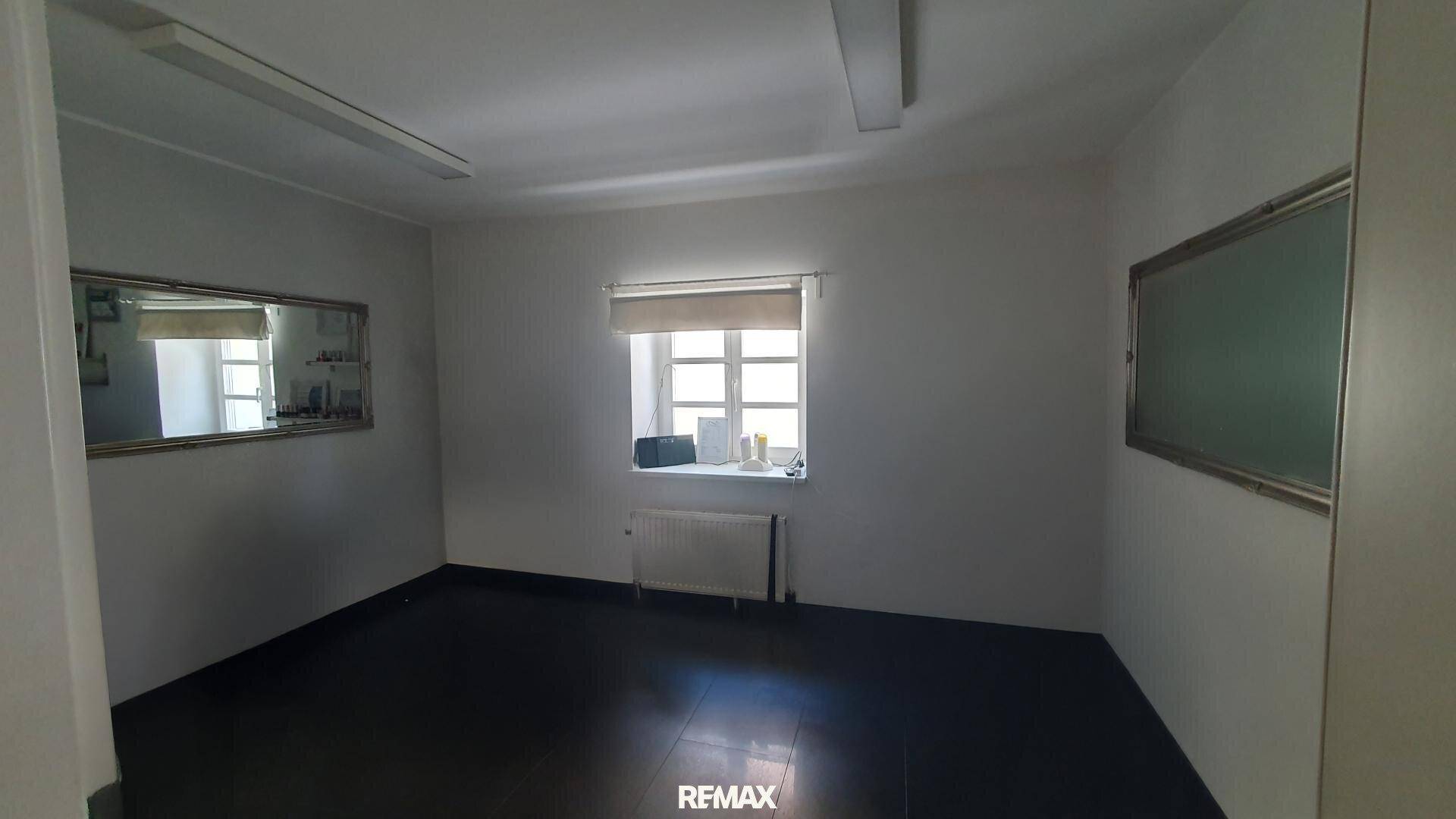 Leerer Raum mit dunklem Fliesenboden, einem Fenster mit Jalousie und zwei Wandspiegeln, geeignet als Büro.