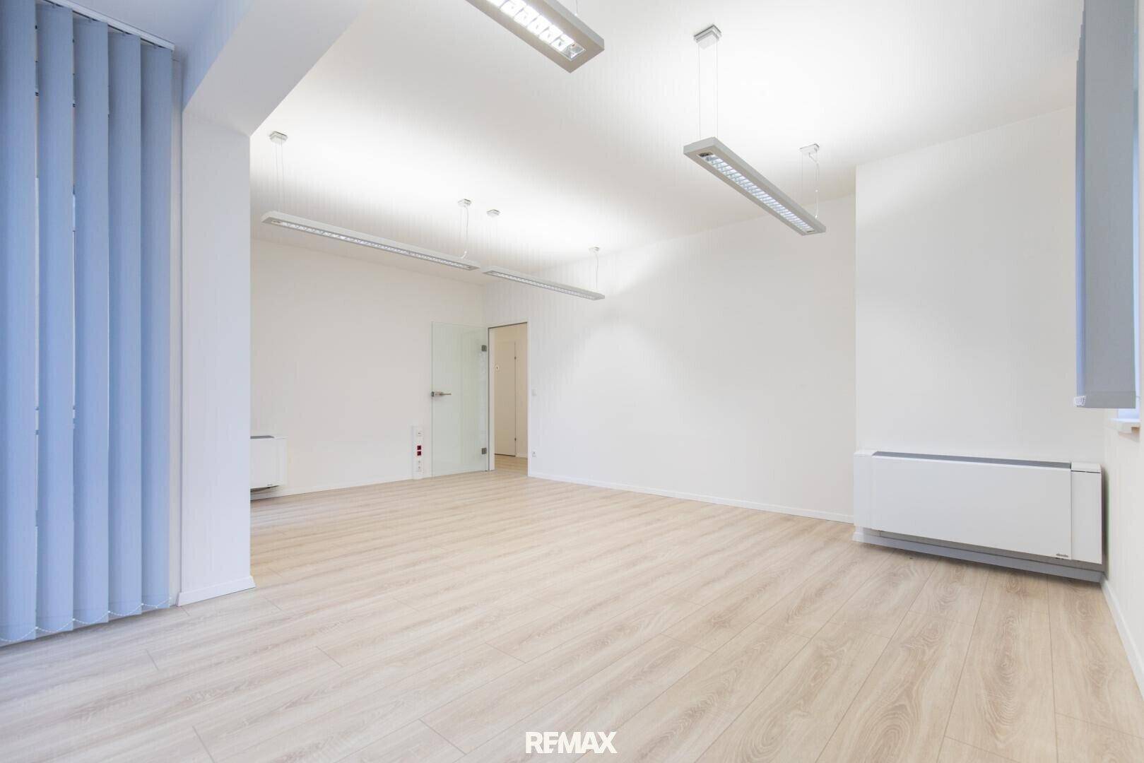 Leerer Büroraum mit hellem Laminatboden, weißen Wänden und einer Tür, die zu einem weiteren Bereich führt.