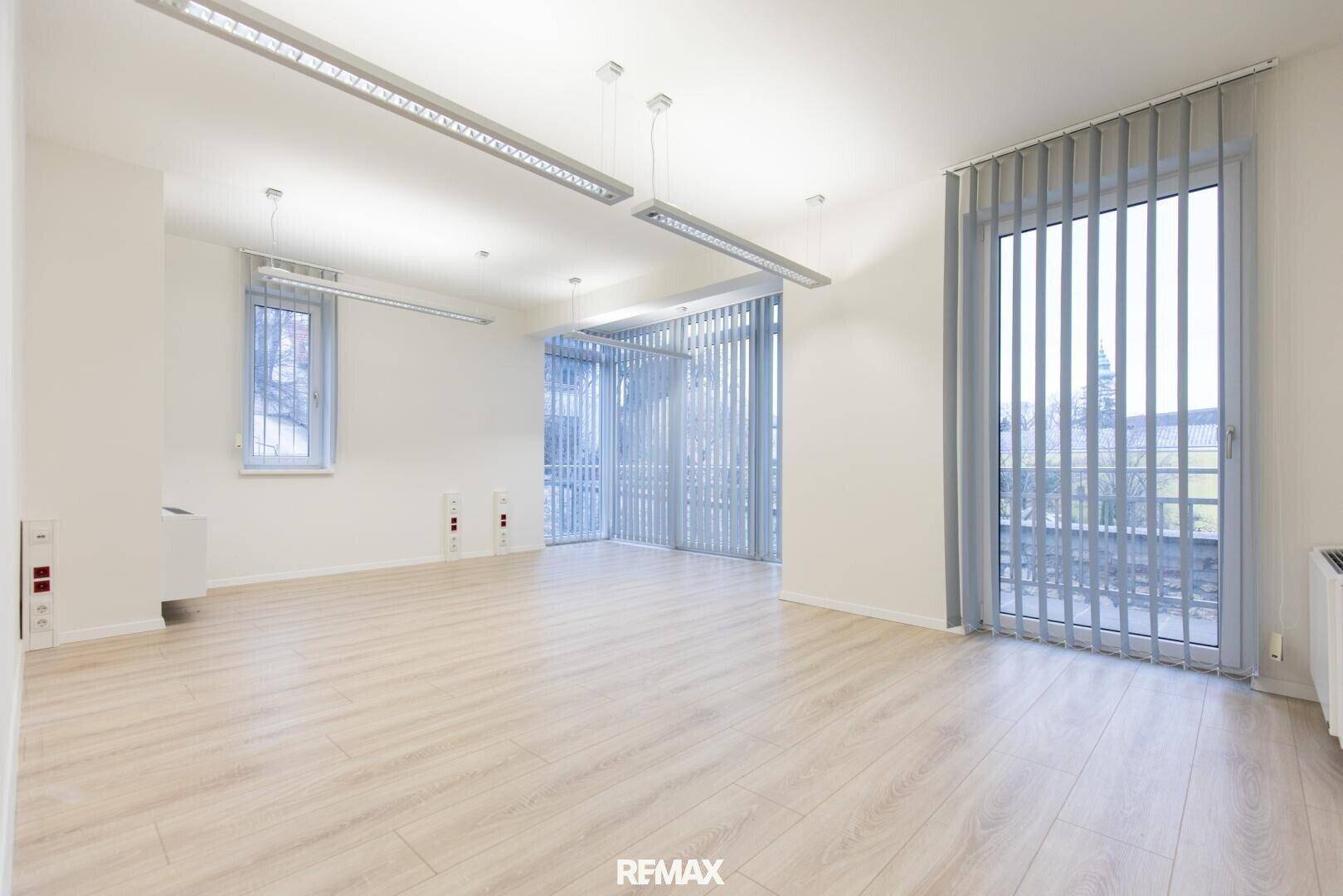 Heller Büroraum mit Laminatboden und drei großen Fenstern, die viel Tageslicht hereinlassen.