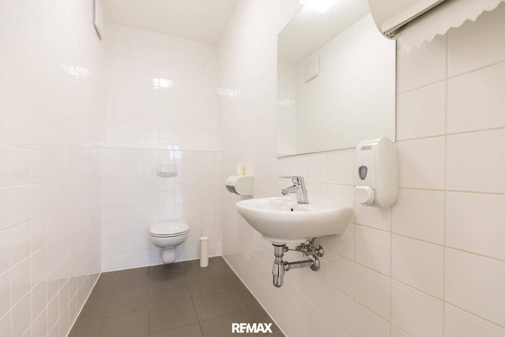 Helles Badezimmer mit weißen Fliesen an den Wänden und dunklen Bodenfliesen, ausgestattet mit WC und Waschbecken.