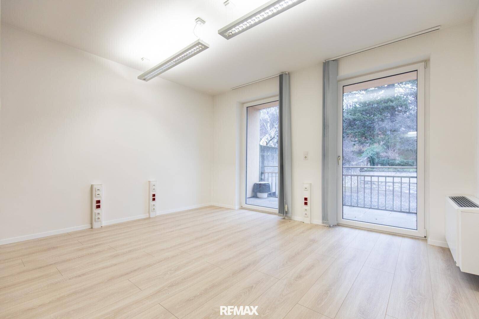 Heller Büroraum mit Laminatboden und zwei großen Glastüren, die Zugang zu einem Außenbereich bieten.