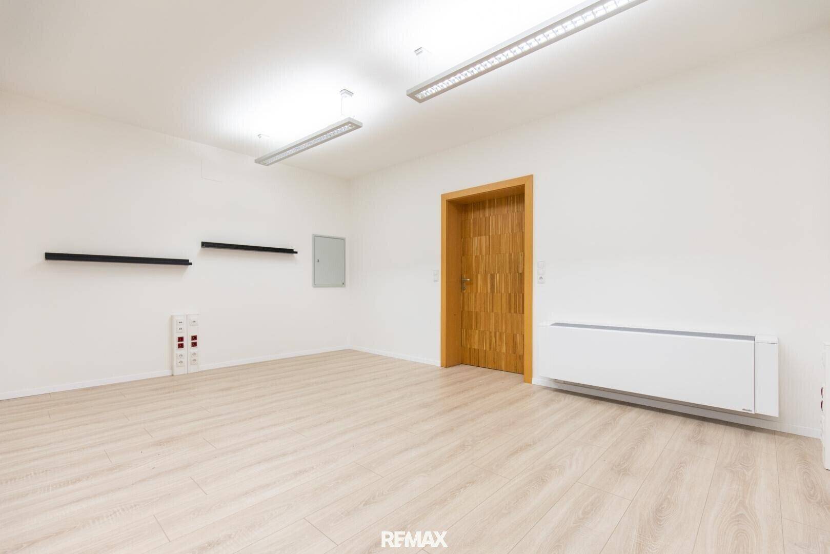 Geräumiger Büroraum mit hellem Laminatboden, weißen Wänden und einer Holztür.