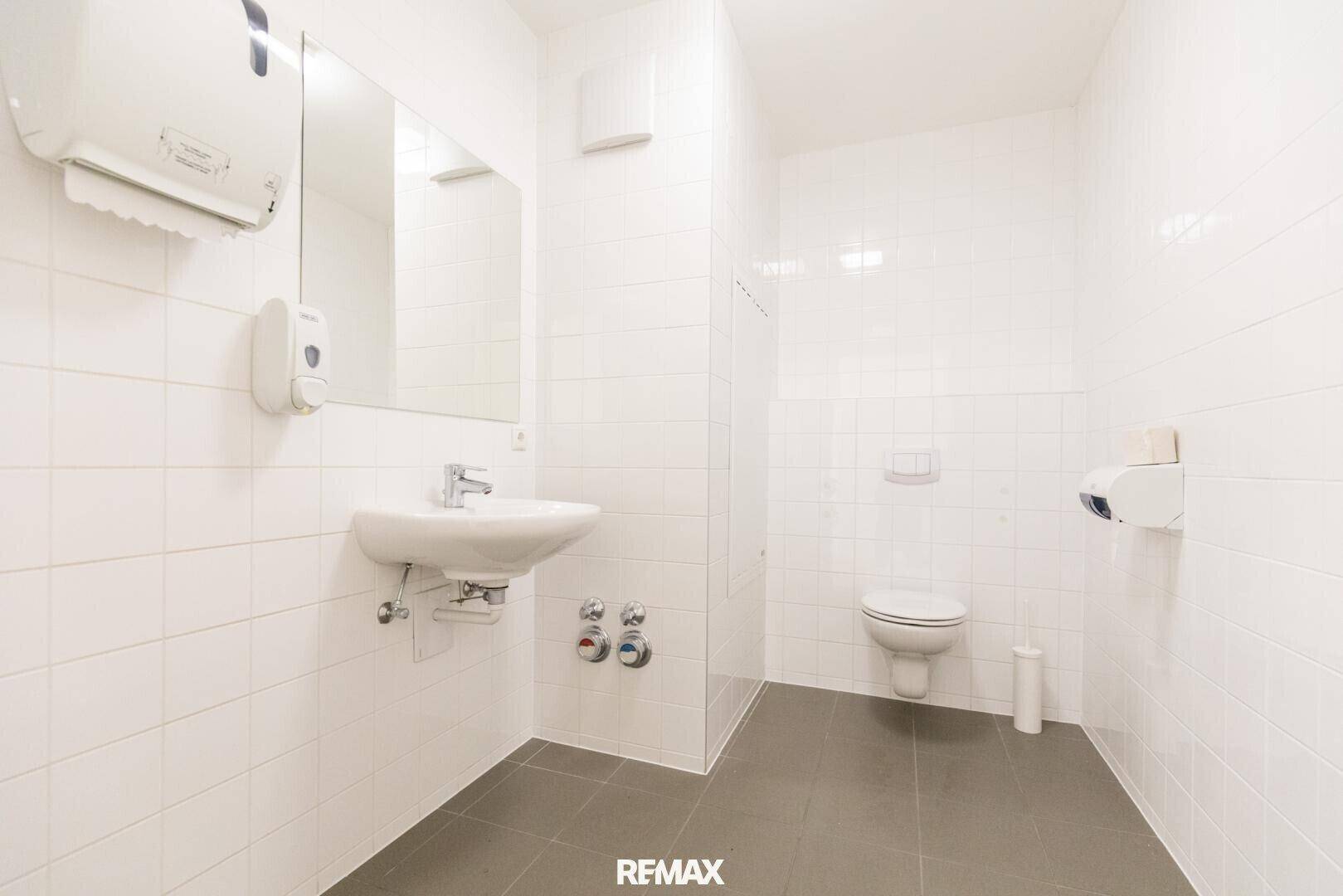 Sauberes Badezimmer mit weißen Wandfliesen und dunklem Fliesenboden, inklusive Waschbecken und Toilette.