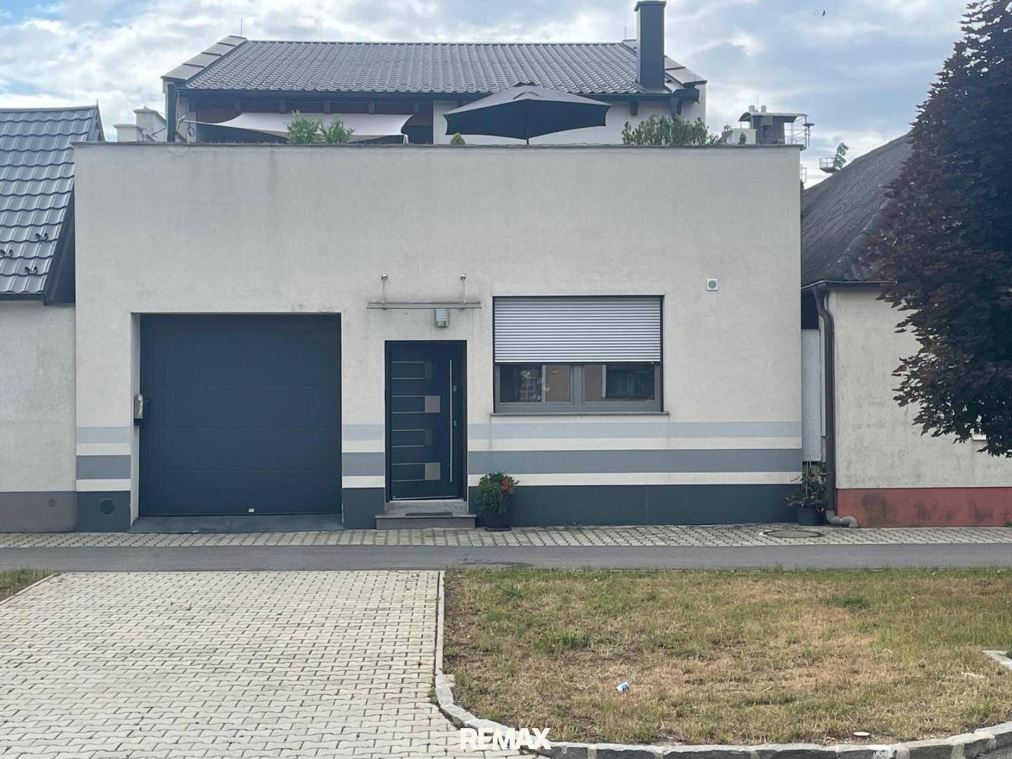 Moderne Gebäudefassade mit Garagentor und Eingangstür, umgeben von einer gepflasterten Auffahrt.