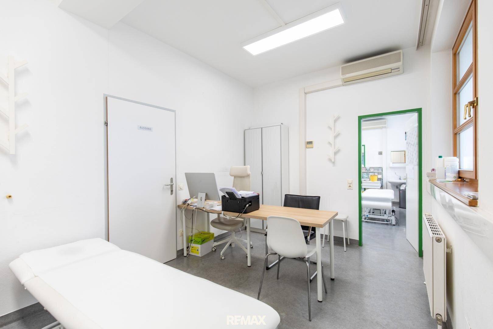 Helles Untersuchungszimmer mit Behandlungsliege, Schreibtisch und Zugang zu einem weiteren Raum, funktional eingerichtet.