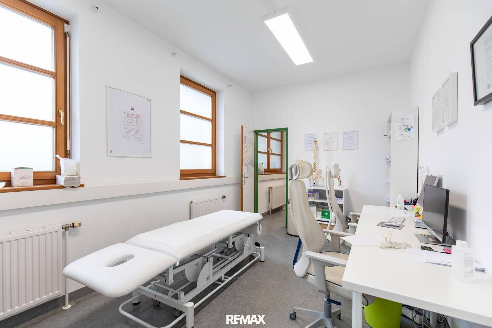 Geräumiges Untersuchungszimmer mit zwei Fenstern, Behandlungsliege und Schreibtisch, hell und sauber gehalten.