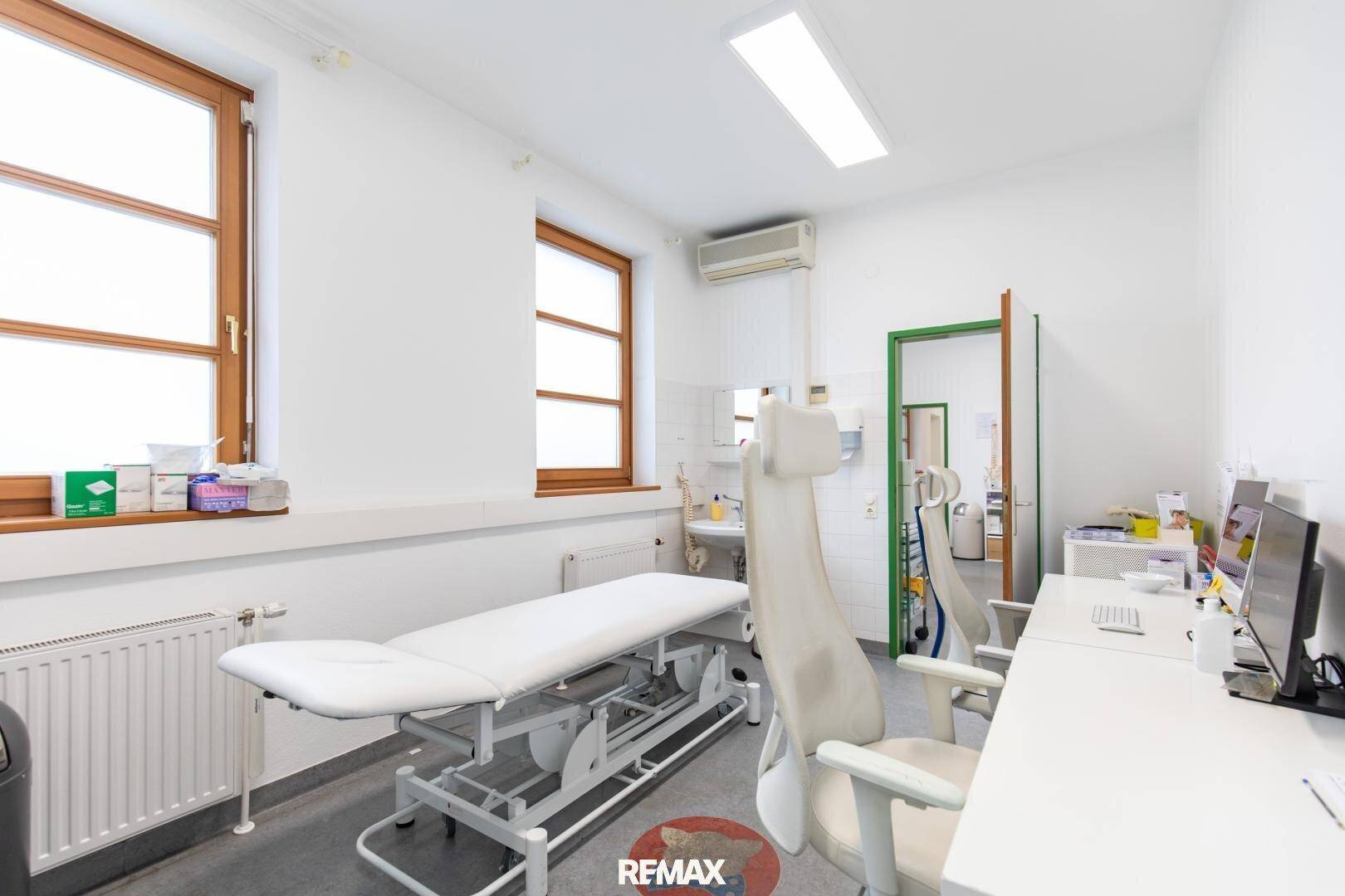 Untersuchungszimmer mit Behandlungsliege, Schreibtisch und Klimaanlage, durch eine Tür verbunden und gut beleuchtet.