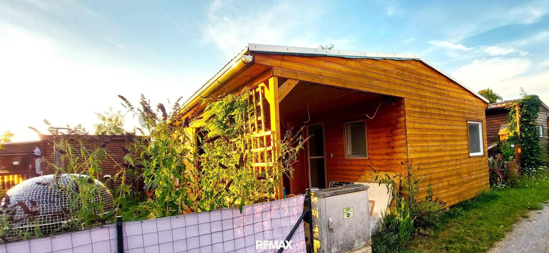 Hölzernes Gartenhaus mit Rankpflanzen und einem Zaun in einer ländlichen Umgebung.