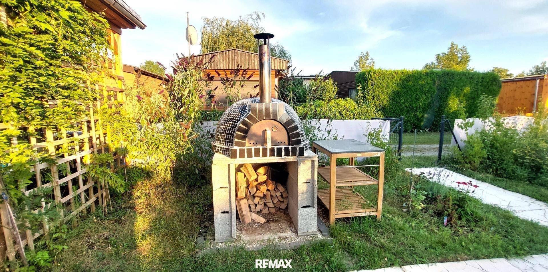 Gemauerter Pizzaofen mit Holzscheiten und einem Beistelltisch im sonnigen Gartenbereich.
