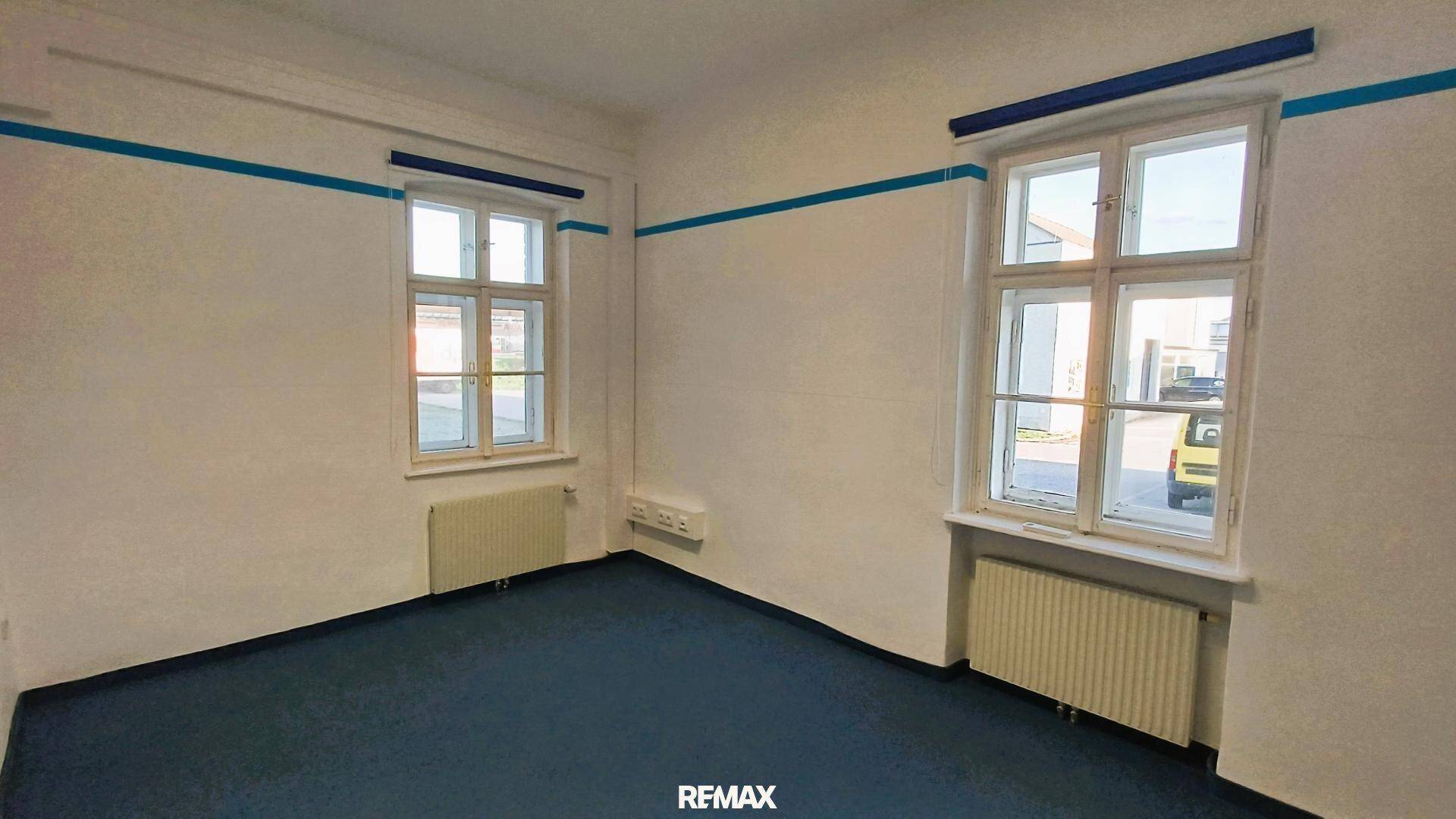 Geräumiges Büro mit blauem Teppichboden, weißen Wänden und zwei großen Fenstern.