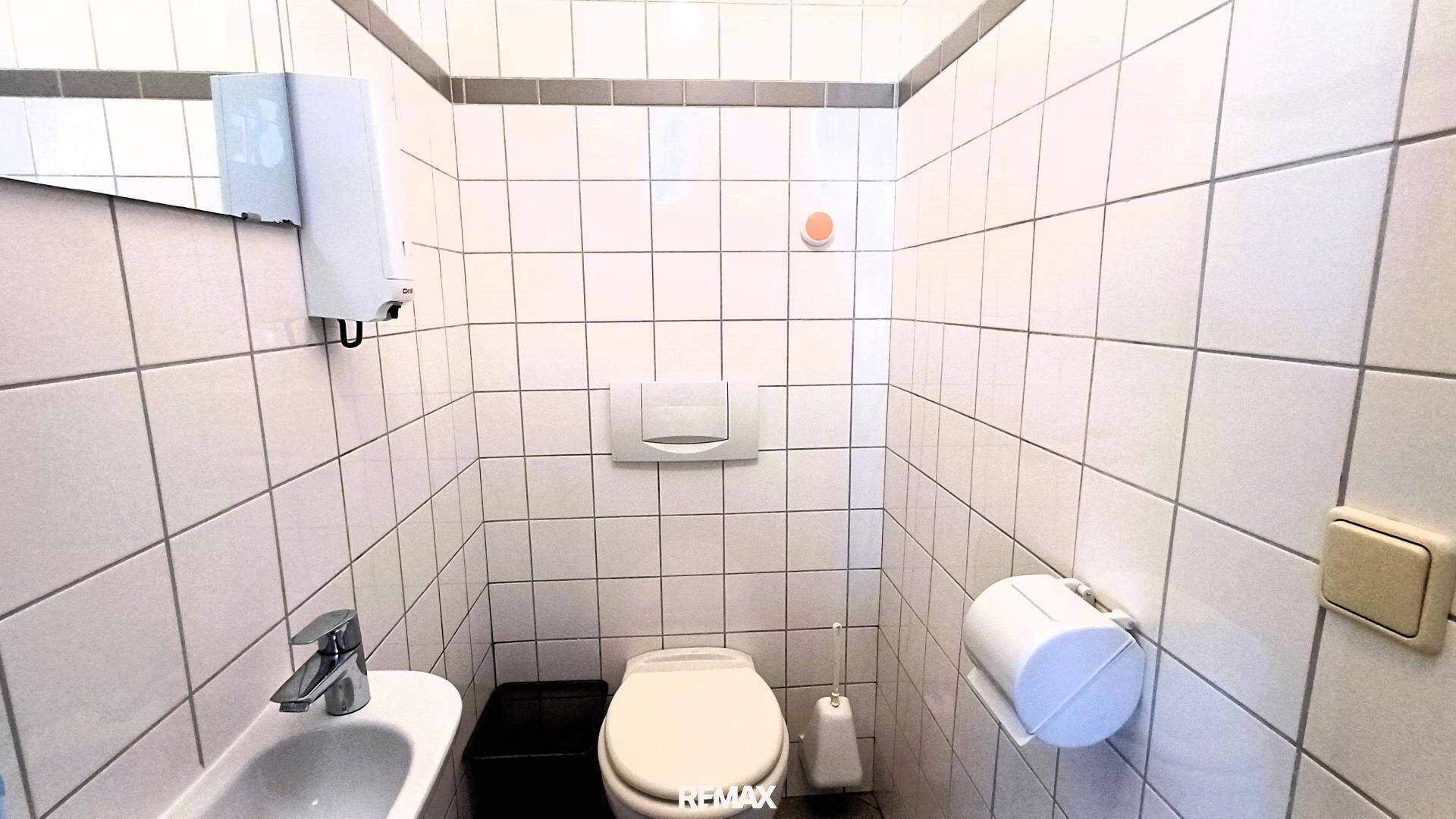 Kleiner Toilettenraum mit weißen Fliesen, einem Waschbecken und einer Wandtoilette.