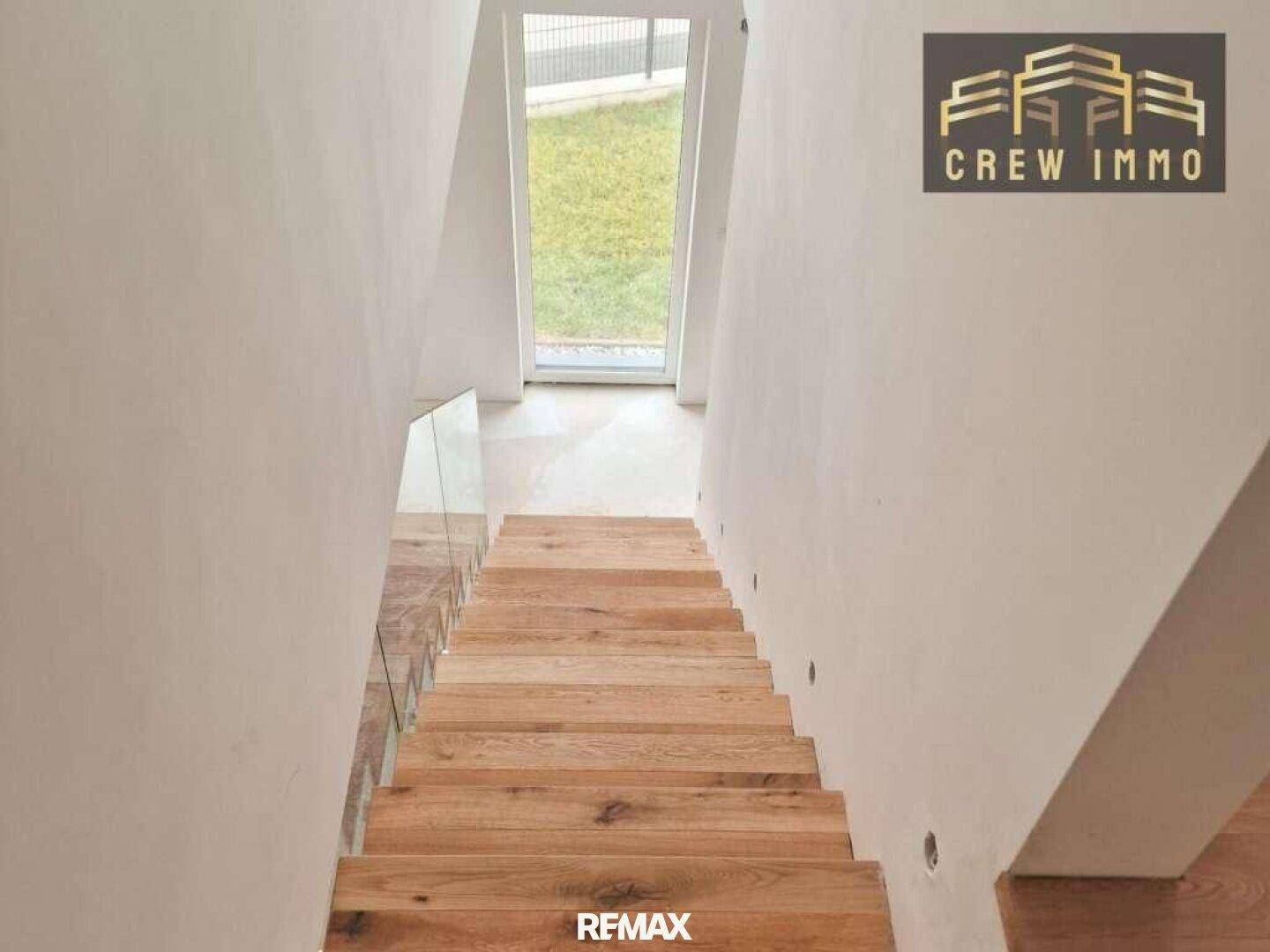 Blick von oben auf eine moderne Holztreppe mit Glasgeländer, die zu einem Ausgang führt.