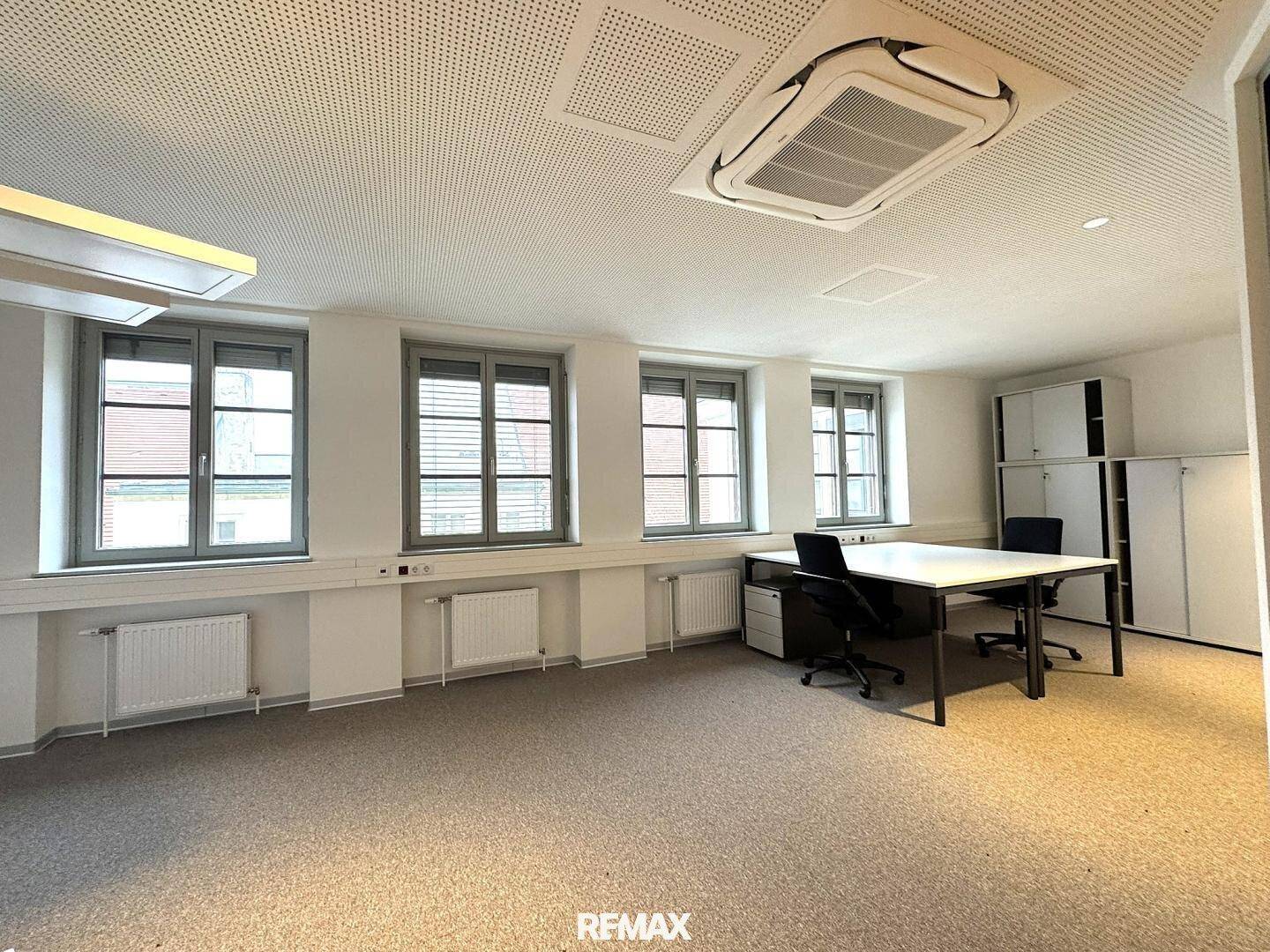 Büroraum mit Teppichboden, Fenstern mit Jalousien und integrierten Schränken.