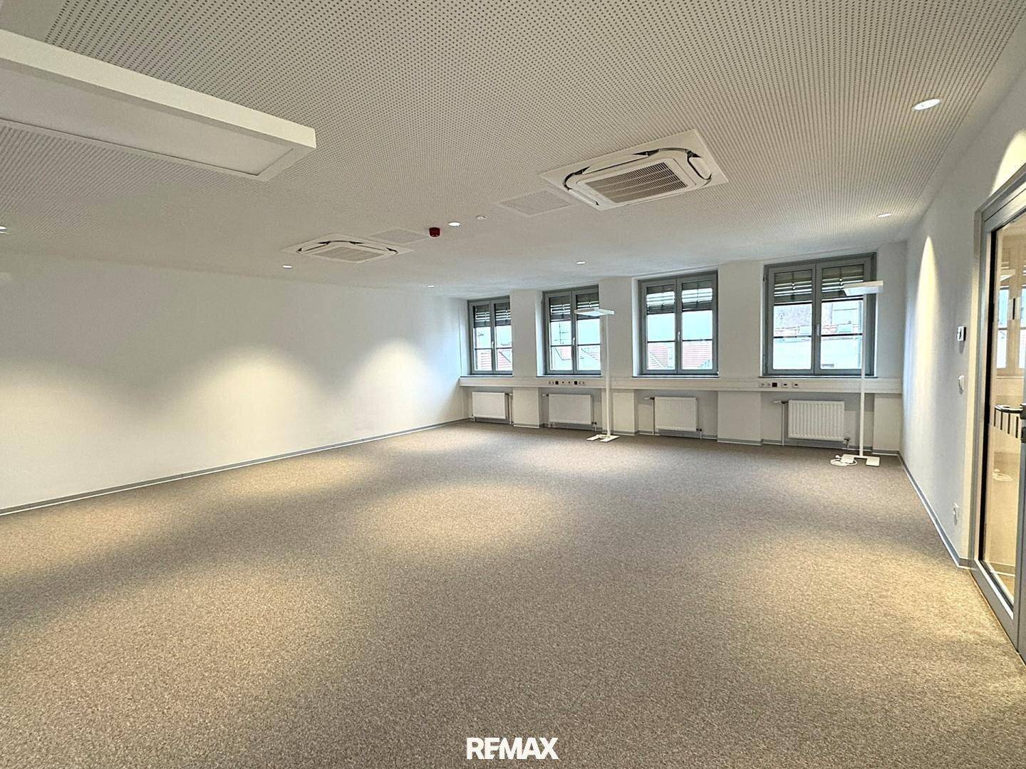 Heller Büroraum mit Teppichboden, großen Fenstern und Klimaanlagen an der Decke.