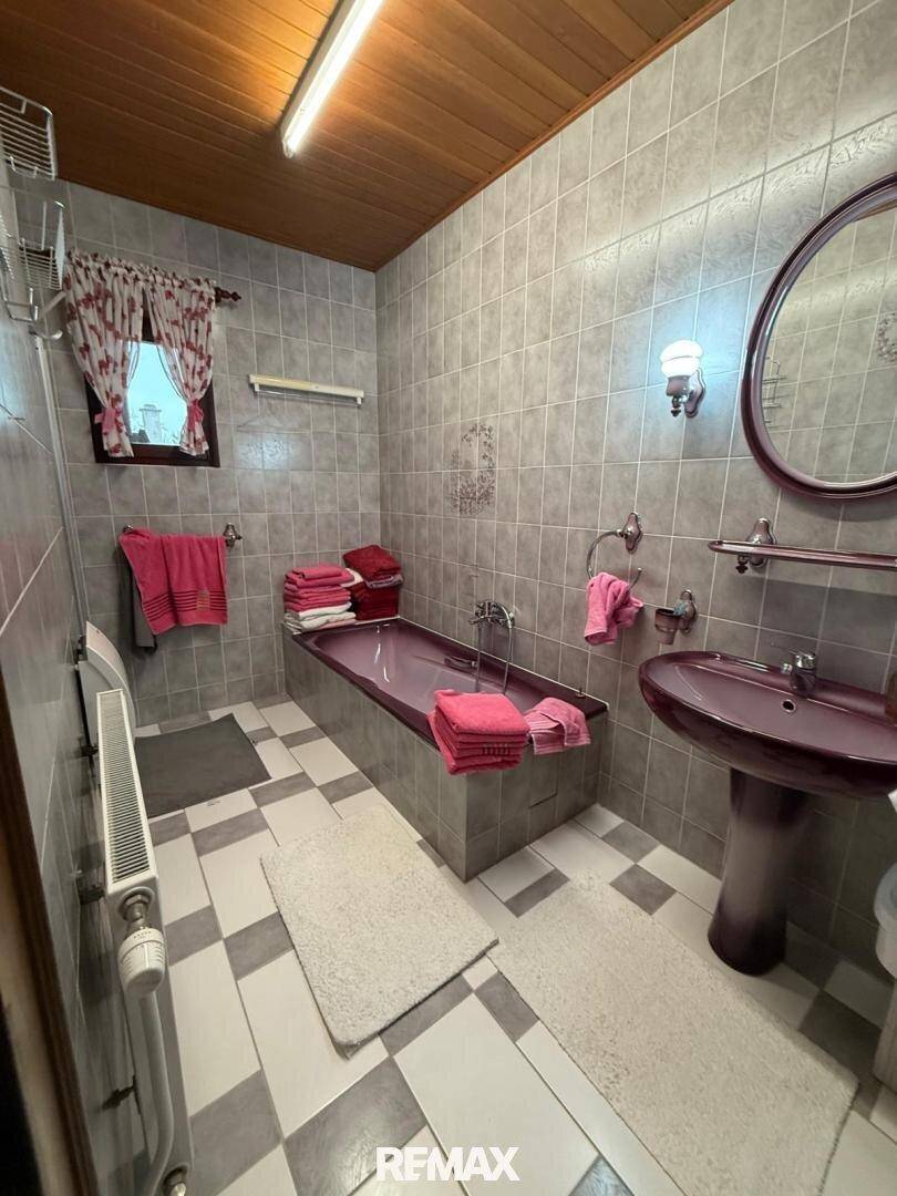 Gefliestes Badezimmer in Grautönen mit Badewanne, Waschbecken und Fenster im 80er-Jahre-Stil.