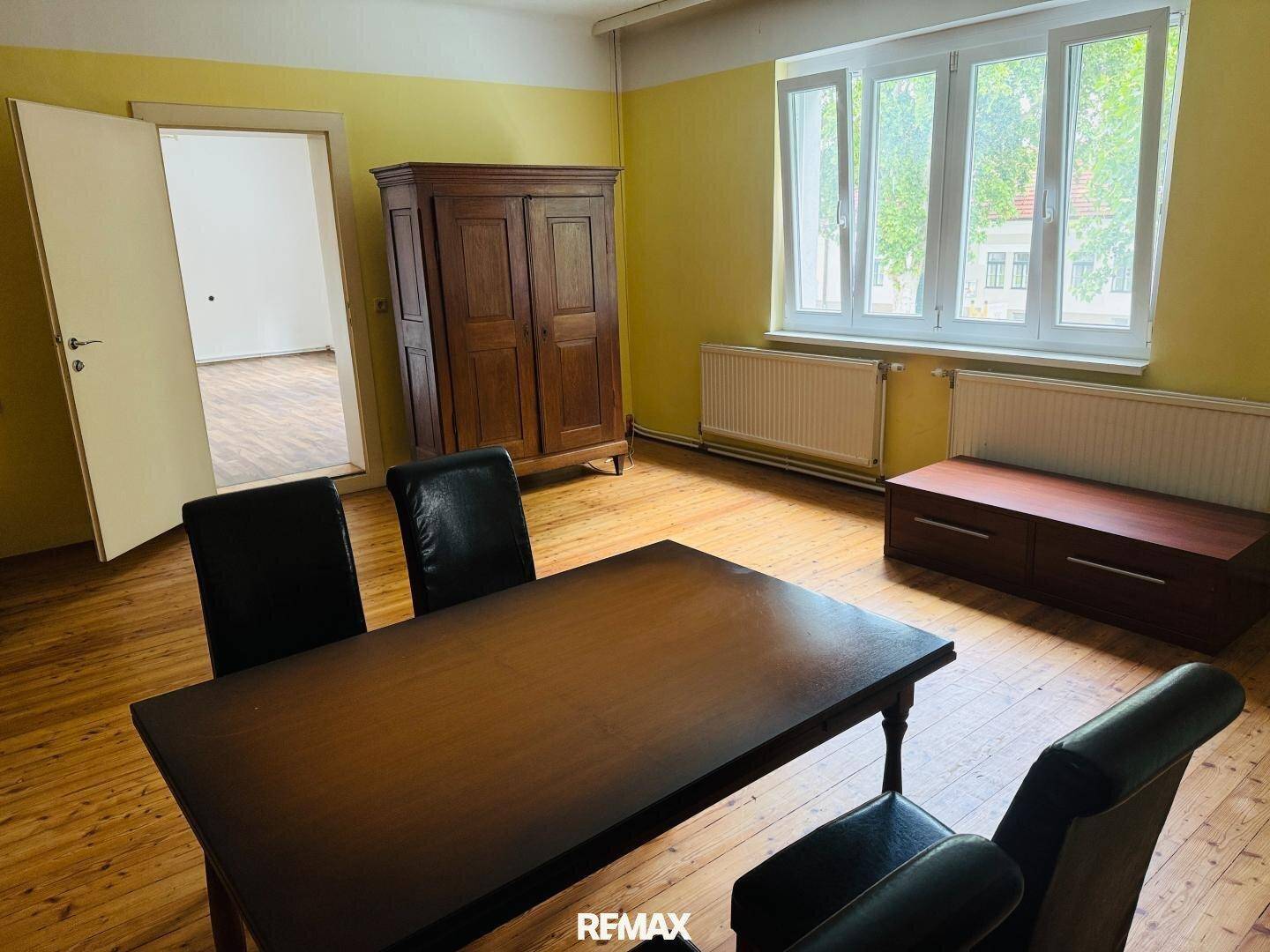 Geräumiges Büro mit Holzboden, gelben Wänden und einem großen Fenster, möbliert mit Tisch und Stühlen.