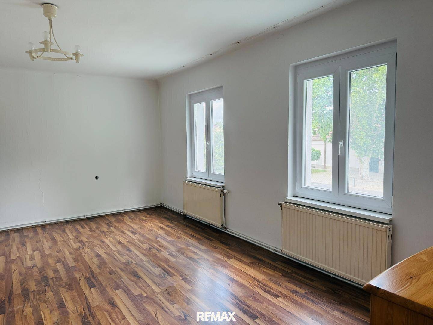 Heller Wohnraum mit Holzboden, weißen Wänden und zwei großen Fenstern mit Blick nach draußen.