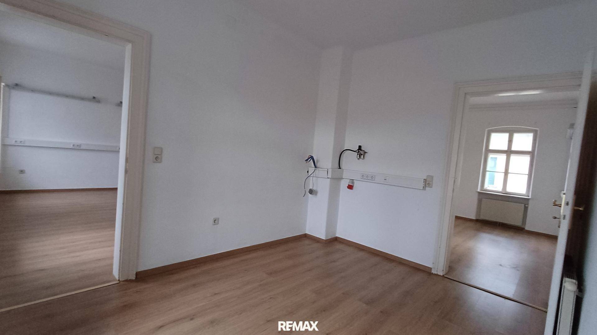 Heller Büroraum mit Laminatboden und zwei offenen Durchgängen zu angrenzenden Zimmern, mit Wandinstallationen.