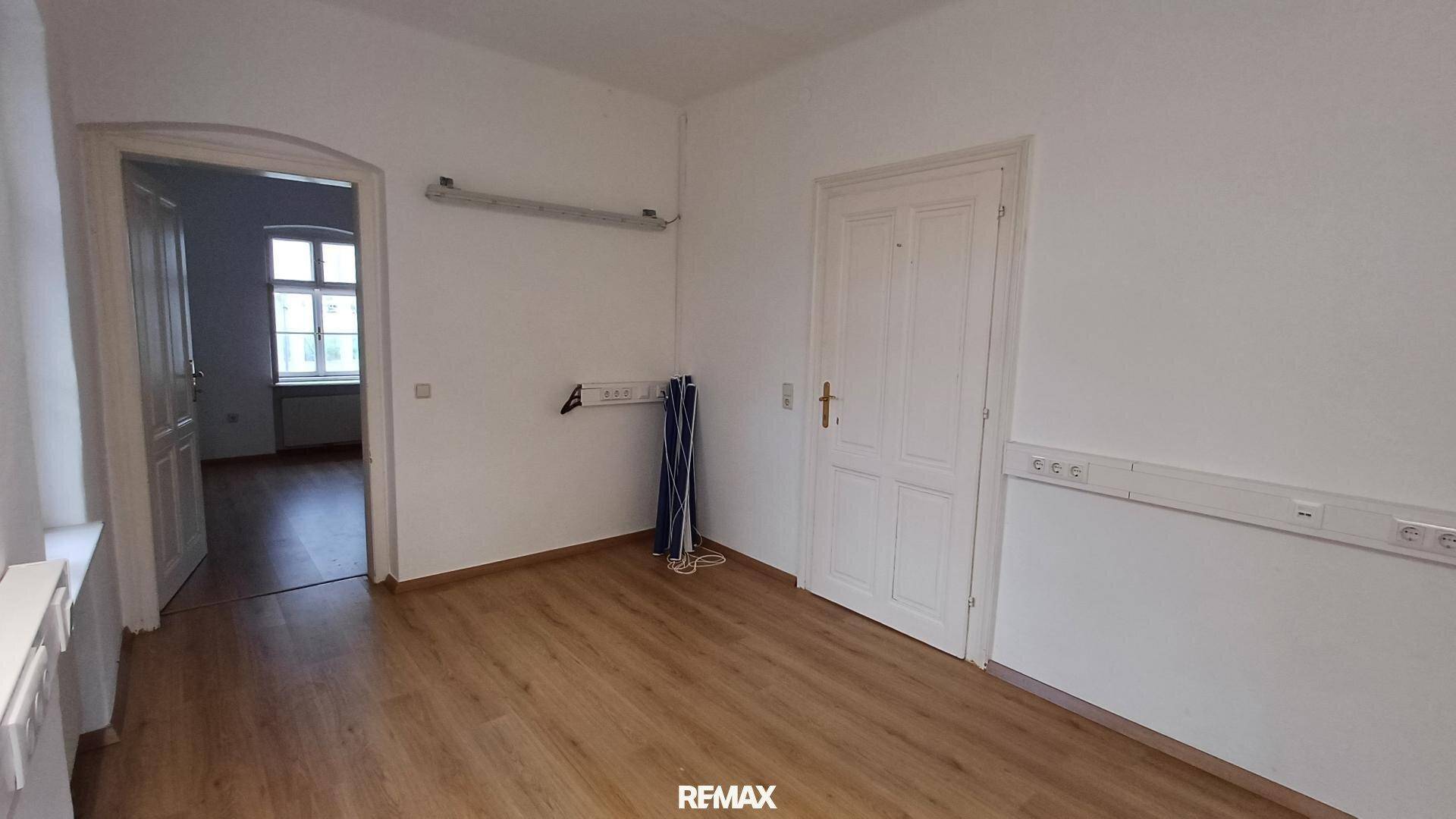 Geräumiger Büroraum mit Laminatboden und Zugang zu einem weiteren Zimmer, ausgestattet mit Steckdosenleisten.