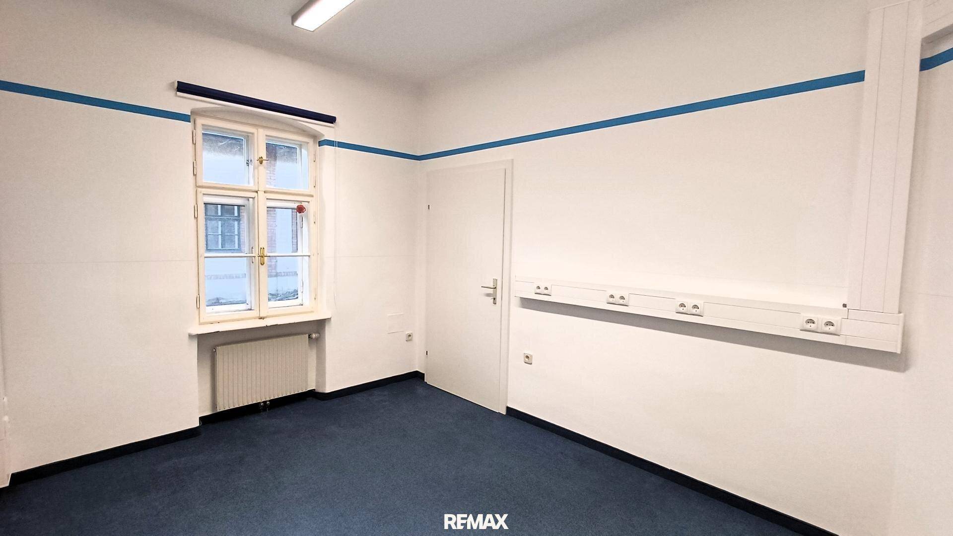 Heller Raum mit blauem Teppichboden, weißen Wänden und einem Fenster, das viel Tageslicht spendet.