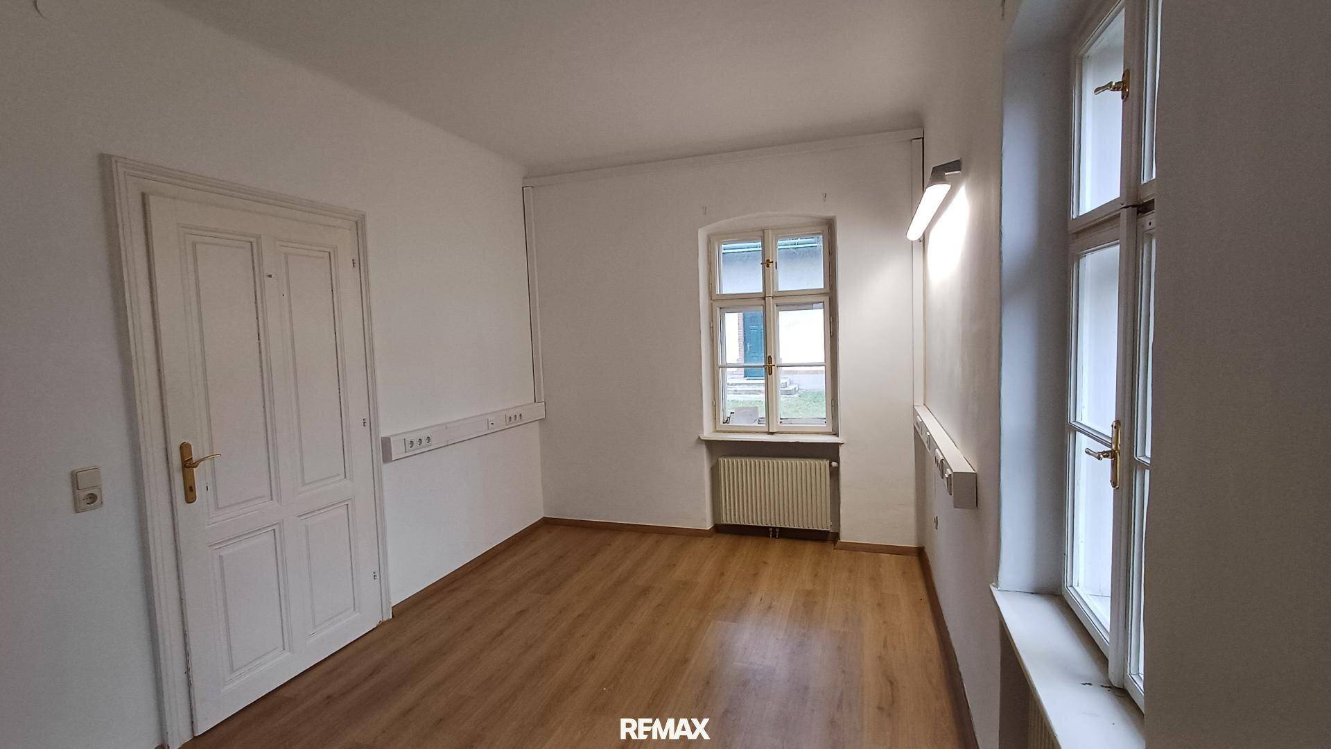 Heller Raum mit Holzoptikboden, weißen Wänden und zwei Fenstern, ideal als Büro oder Arbeitszimmer.
