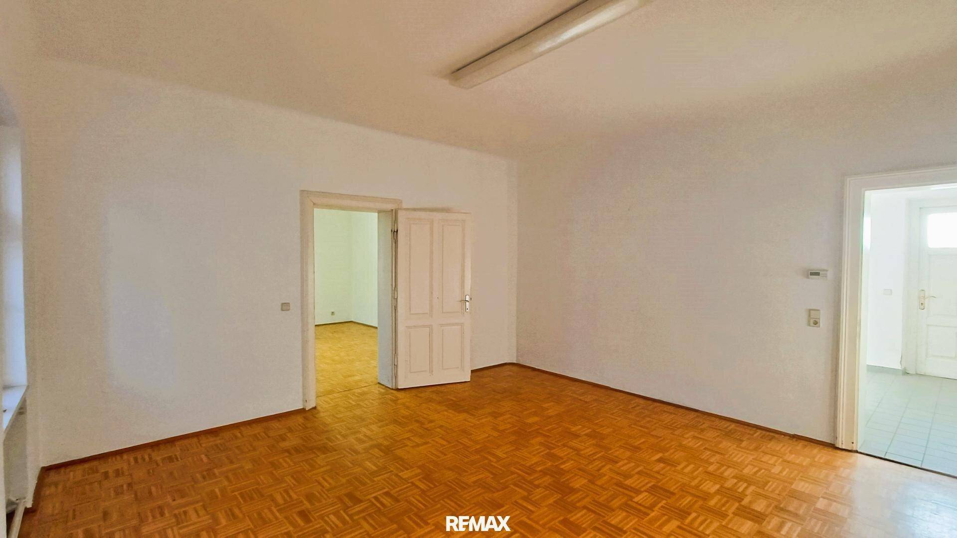 Großer, heller Raum mit Fischgrätparkettboden und weißen Wänden, der durch Doppeltüren zugänglich ist.