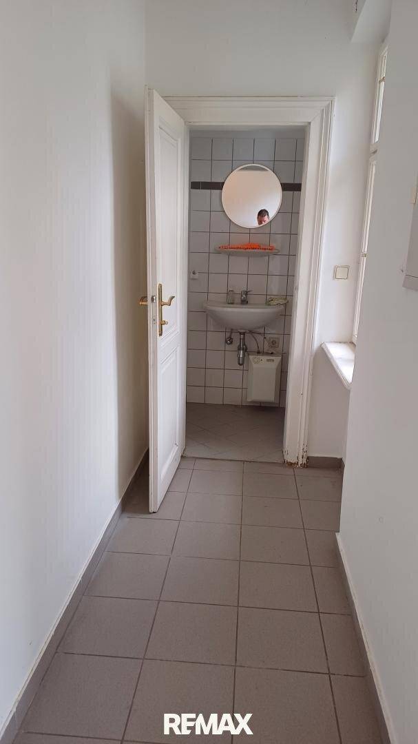 Flur mit grauen Fliesen führt zu einem Badezimmer mit Waschbecken und Spiegel.