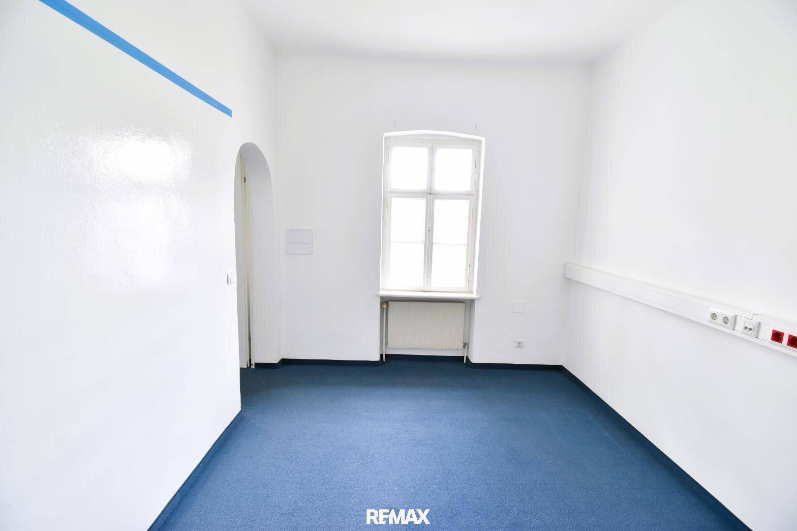 Heller Raum mit blauem Teppichboden, weißen Wänden und einem Fenster, das viel Tageslicht spendet.
