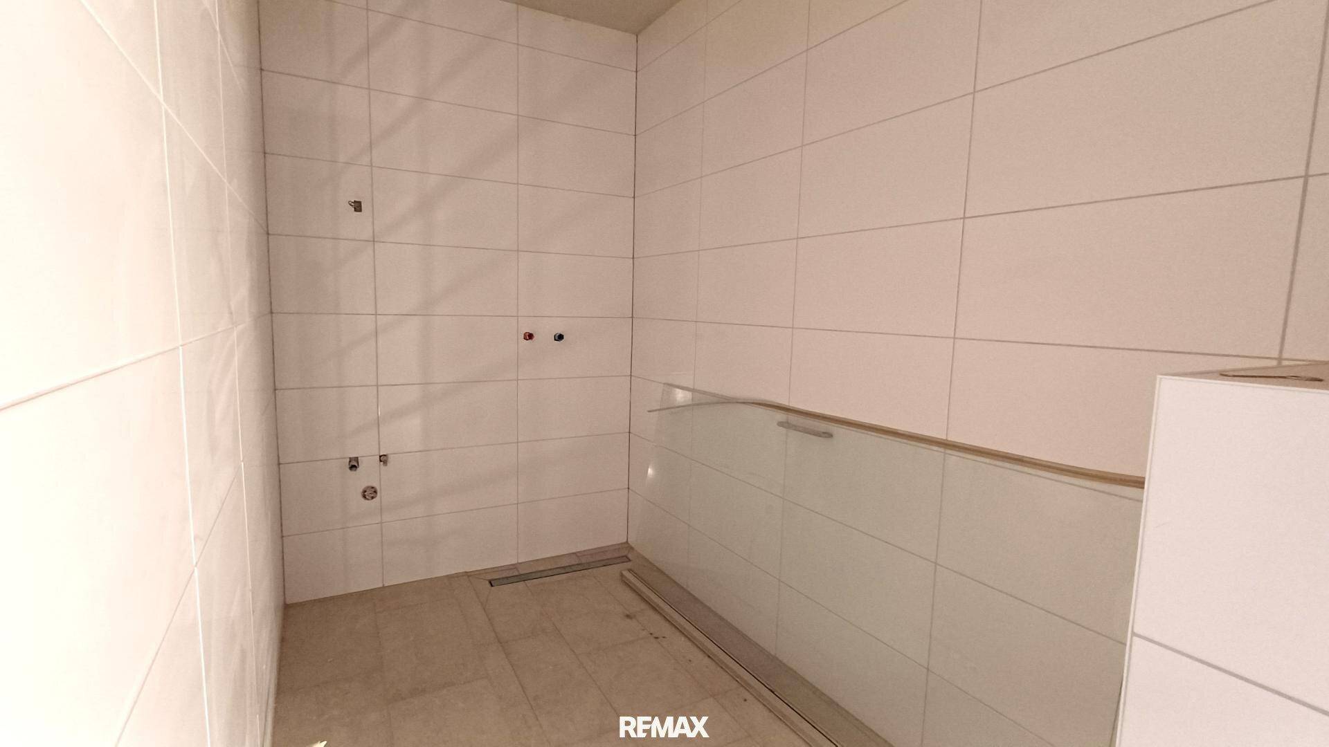 Badezimmer im Rohbau mit weißen Wandfliesen und vorbereitetem Duschbereich.