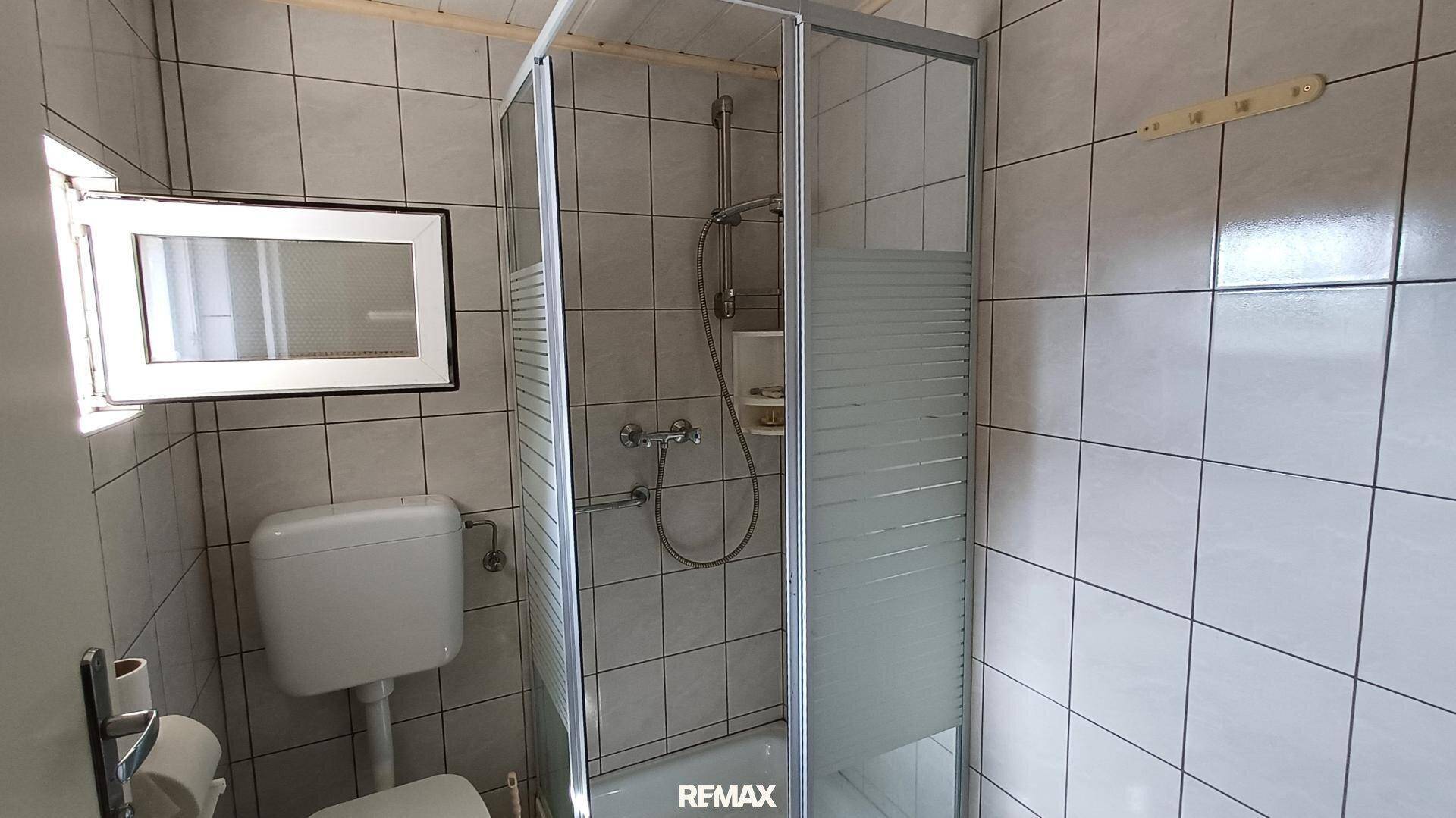 Kleines Badezimmer mit Duschkabine, Toilette und Fenster, komplett gefliest.