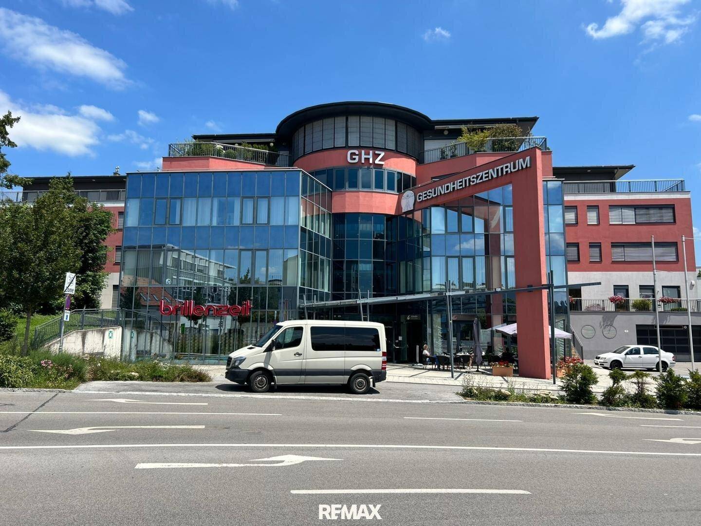 Modernes Gesundheitszentrum mit Glasfassade und umliegenden Parkplätzen unter blauem Himmel.